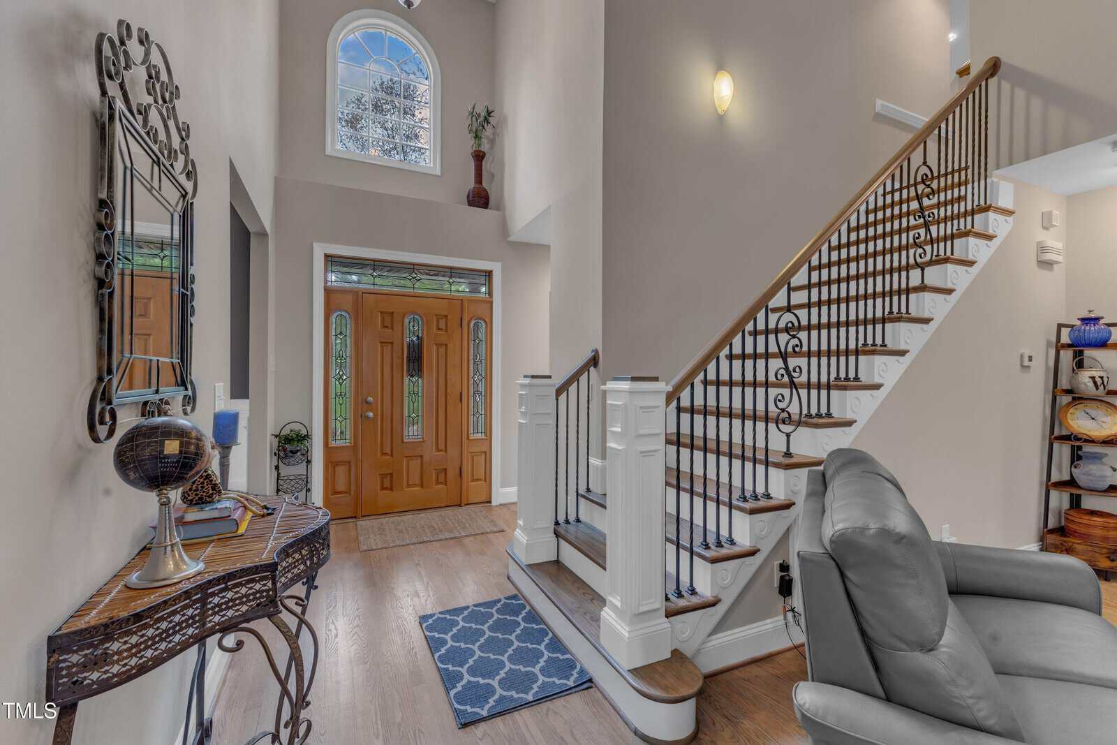 A well-lit foyer with wooden double doors and stained glass windows. A staircase with a wooden and iron railing curves upwards. A decorative table with a mirror is on the left, and a gray armchair is partially visible on the right.
