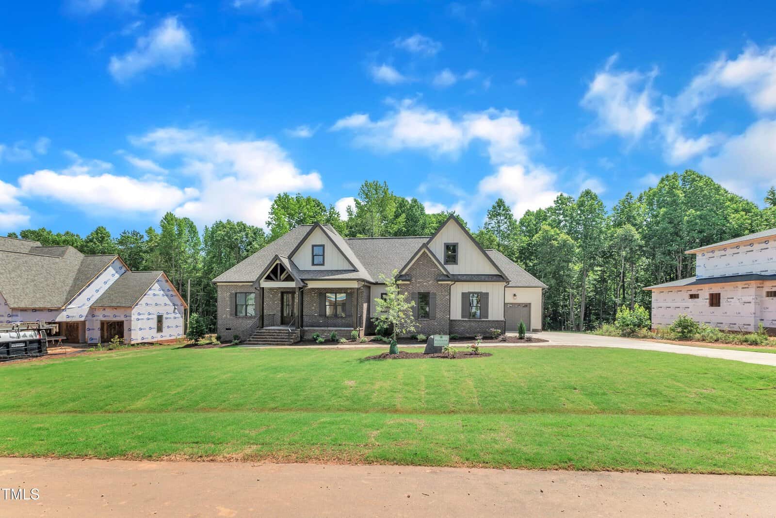 A single-story house with gabled roof, brick and siding exterior, small front porch, and landscaped yard sits between two houses under construction, with trees and blue sky in the background.