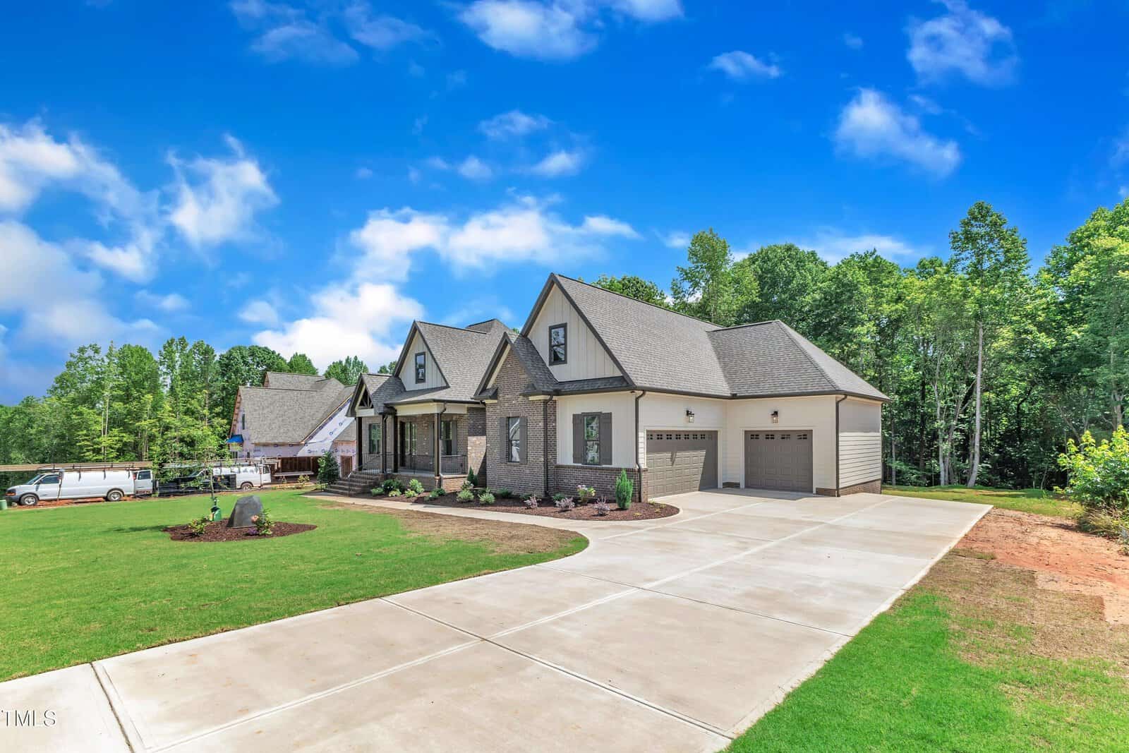 A large, modern suburban house with brick and siding exterior, attached two-car garage, manicured lawn, and a wide concrete driveway, surrounded by trees under a bright blue sky with scattered clouds.