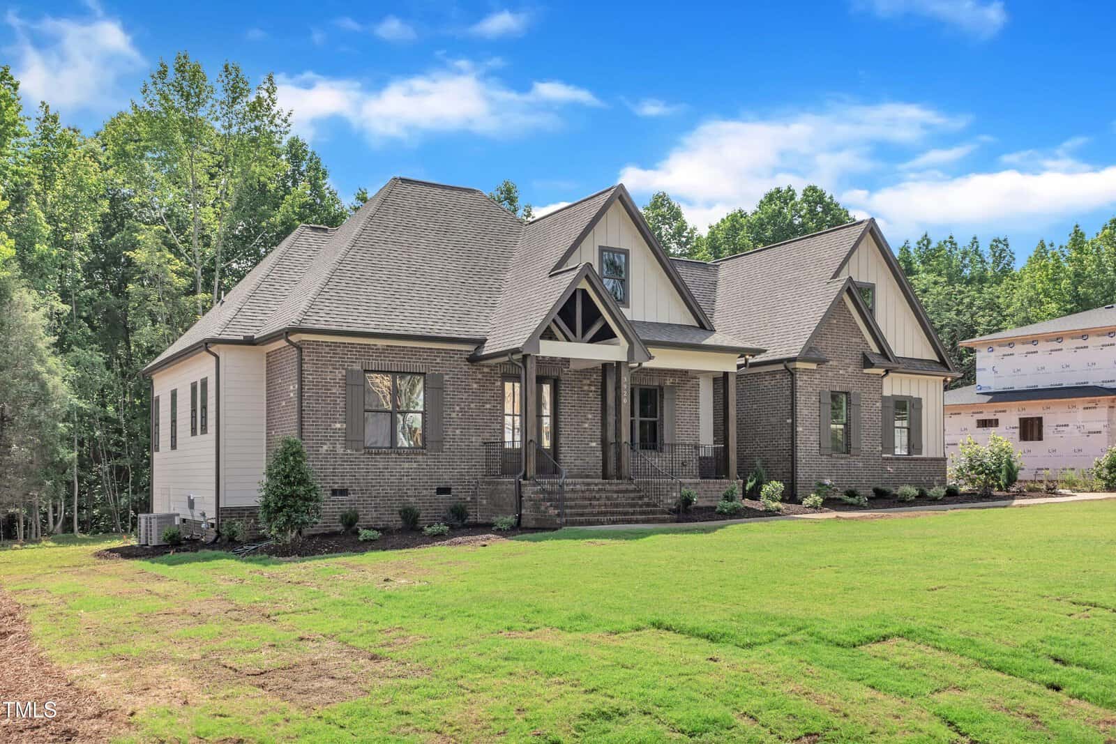 A two-story house with gray brick and light siding exterior, gabled roof, covered front porch with columns, and a newly planted lawn, set against a backdrop of trees and blue sky.