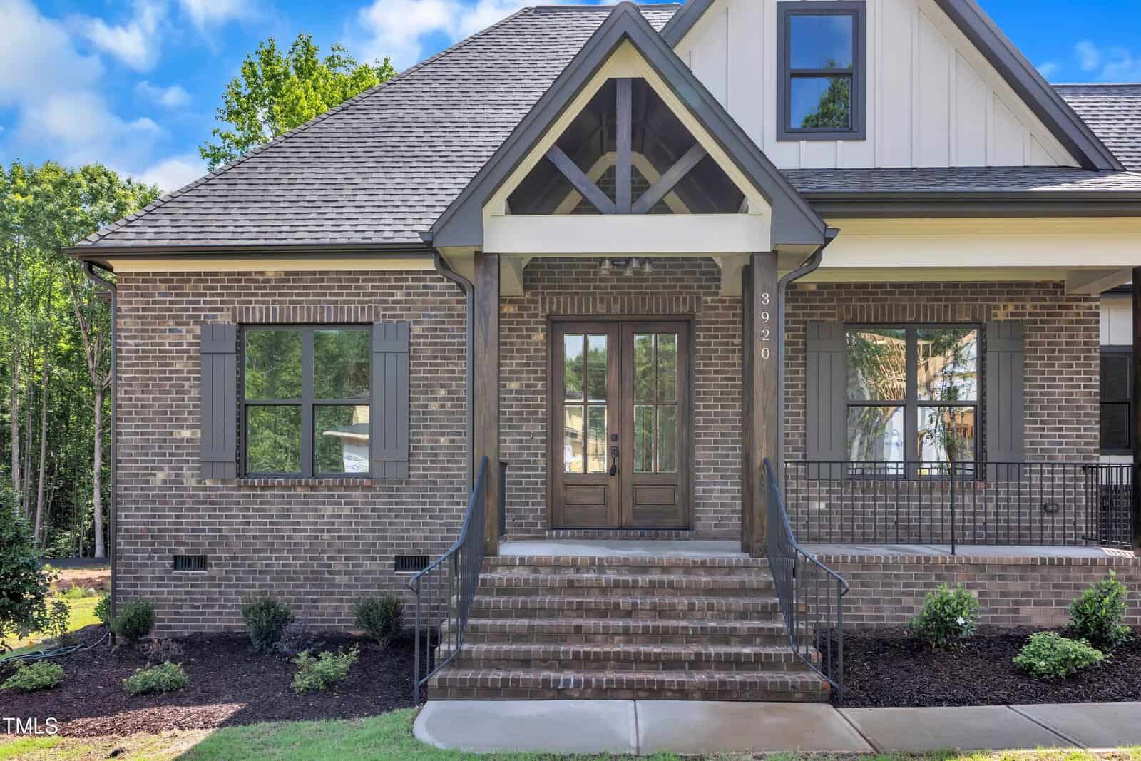 A brick house with a gabled roof has a double wooden front door, steps with metal railings leading to the entrance, two large front windows, and small shrubs along the foundation.