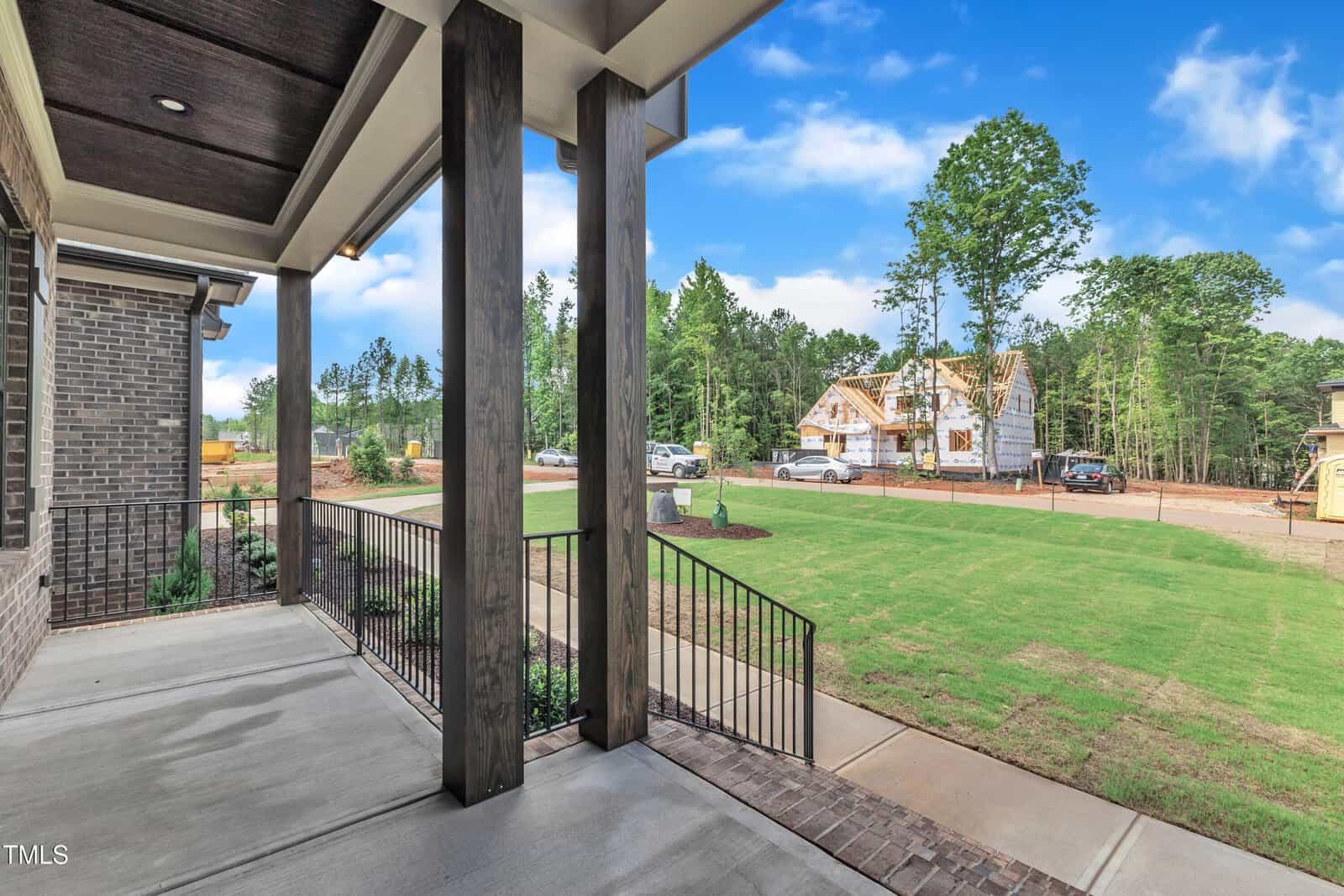 View from a covered front porch with black railings, looking out onto a lawn and a street with houses under construction and several parked vehicles, on a sunny day with a blue sky and scattered clouds.