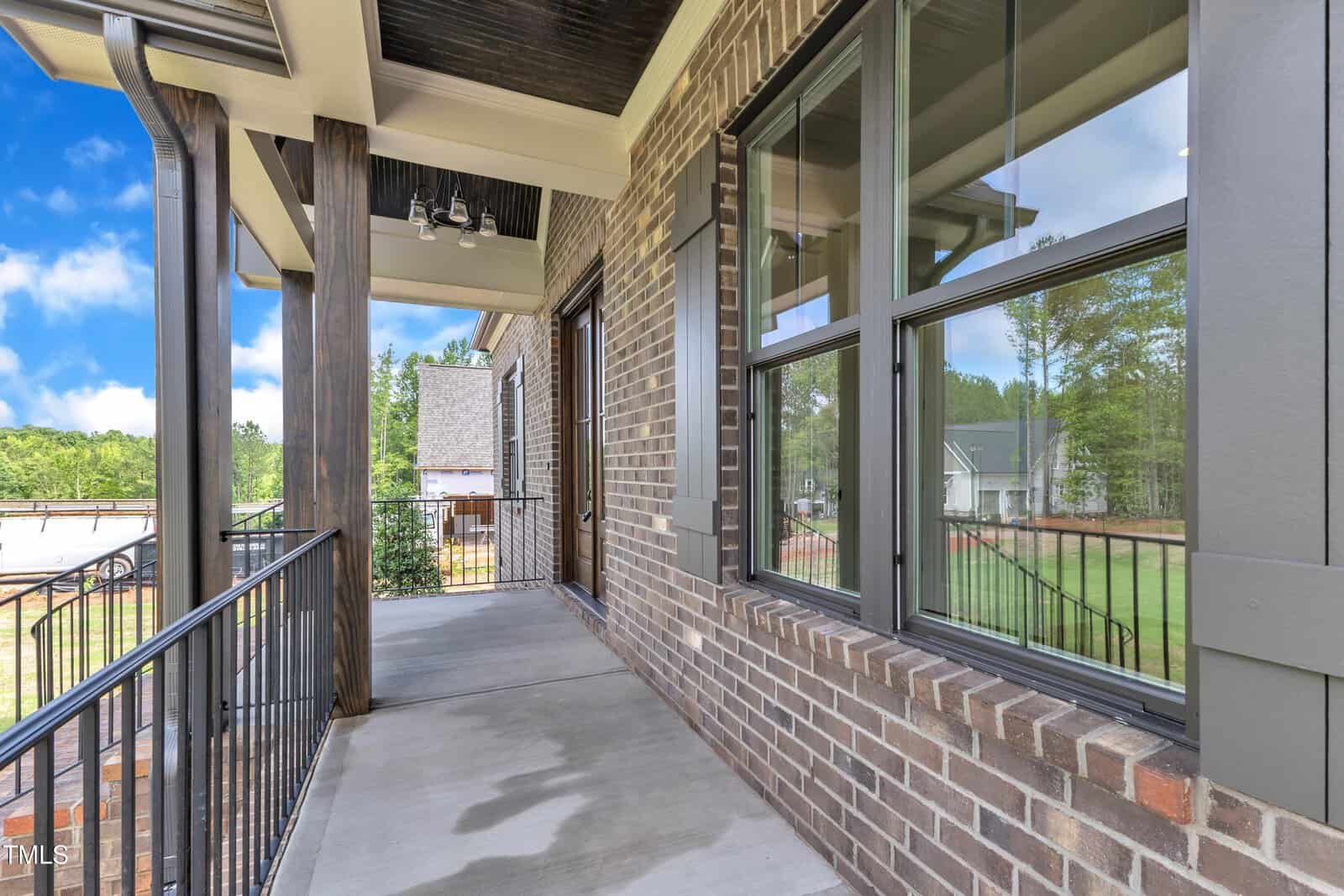 A covered porch with a black metal railing, brick walls, large windows, and a concrete floor. The porch overlooks a grassy yard and nearby houses under a partly cloudy sky.
