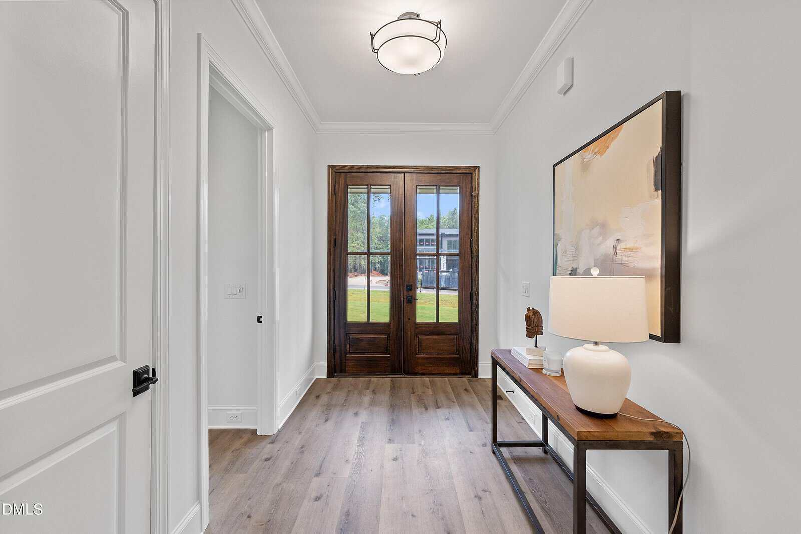 A bright entryway with light-colored walls, wood flooring, and a dark wooden front door with glass panels. A table along the right wall displays a lamp and abstract artwork hangs above it.