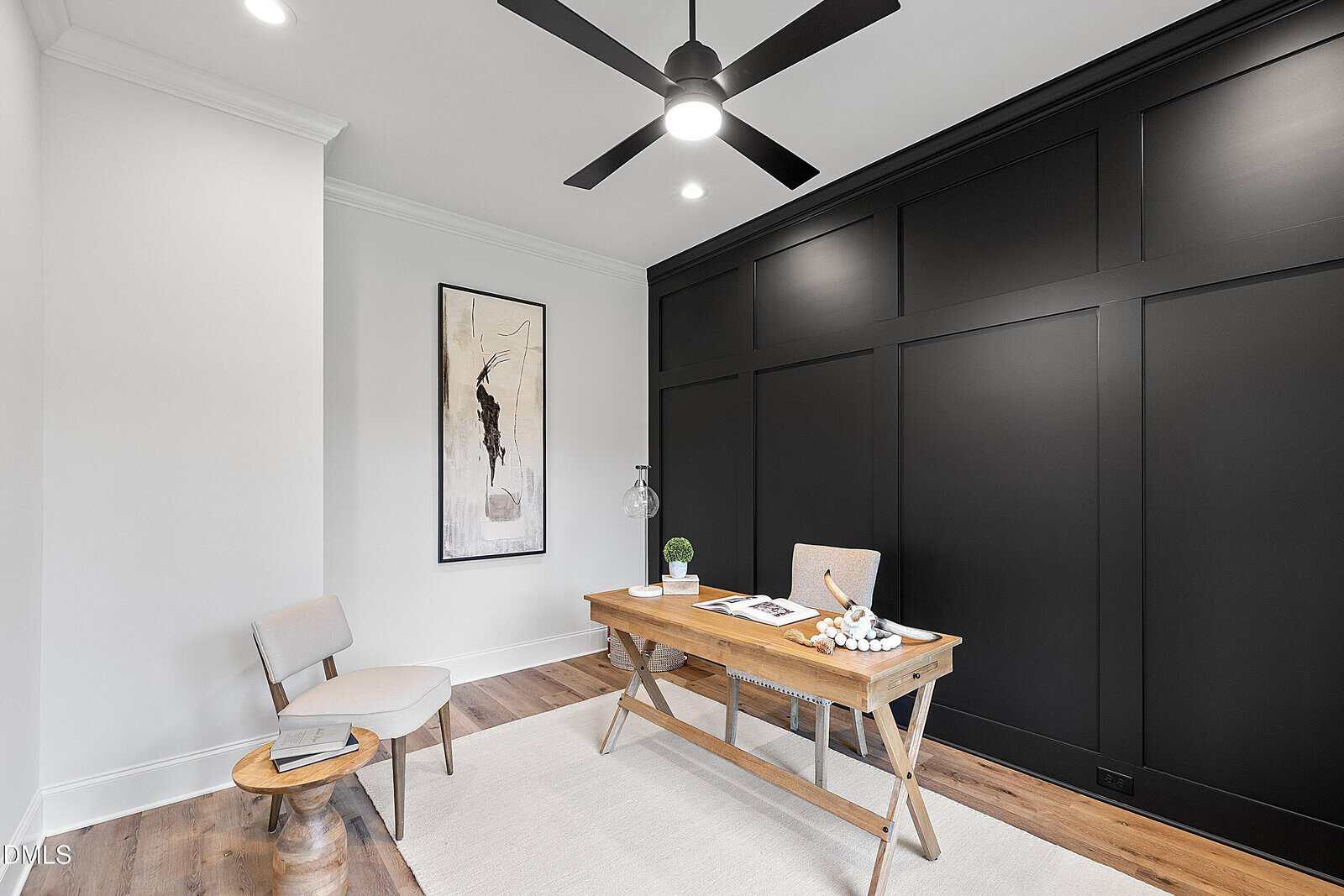 A modern home office features a wooden desk with a chair, a small round side table with a chair, a large abstract art piece on a white wall, a black accent wall, a ceiling fan, and light wood flooring.