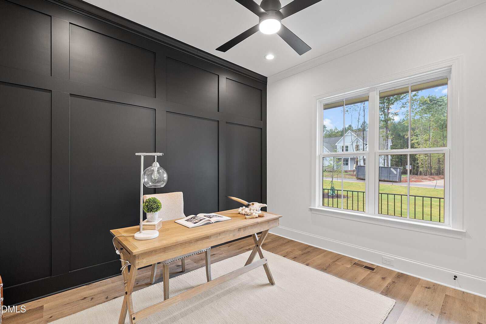 A modern home office with a wooden desk, lamp, plant, and books, set against a black accent wall. A large window lets in natural light and shows a view of houses and trees outside. The floor is light wood with a white rug.