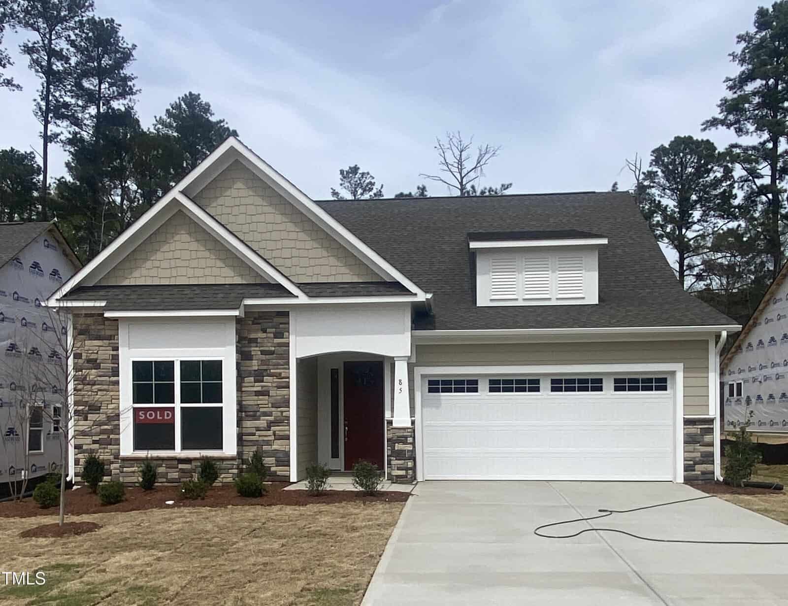 A newly built single-story house with stone and siding exterior, a covered porch, and a double garage. A "SOLD" sign is visible in the front window. The driveway and yard appear freshly completed.