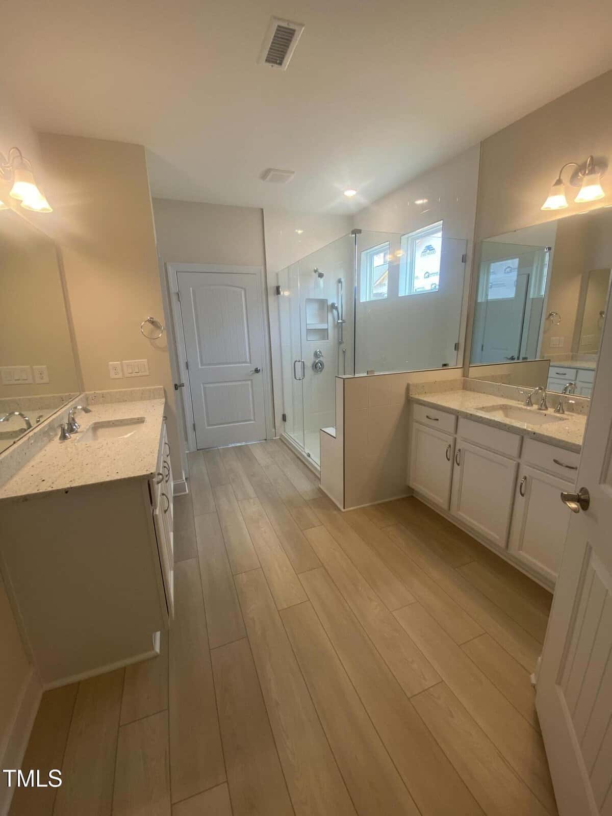 Modern bathroom with light wood floors, double sinks, white cabinets, a large glass-enclosed shower, and beige walls. Multiple mirrors and ceiling lights are visible. A closed door is at the far end of the room.