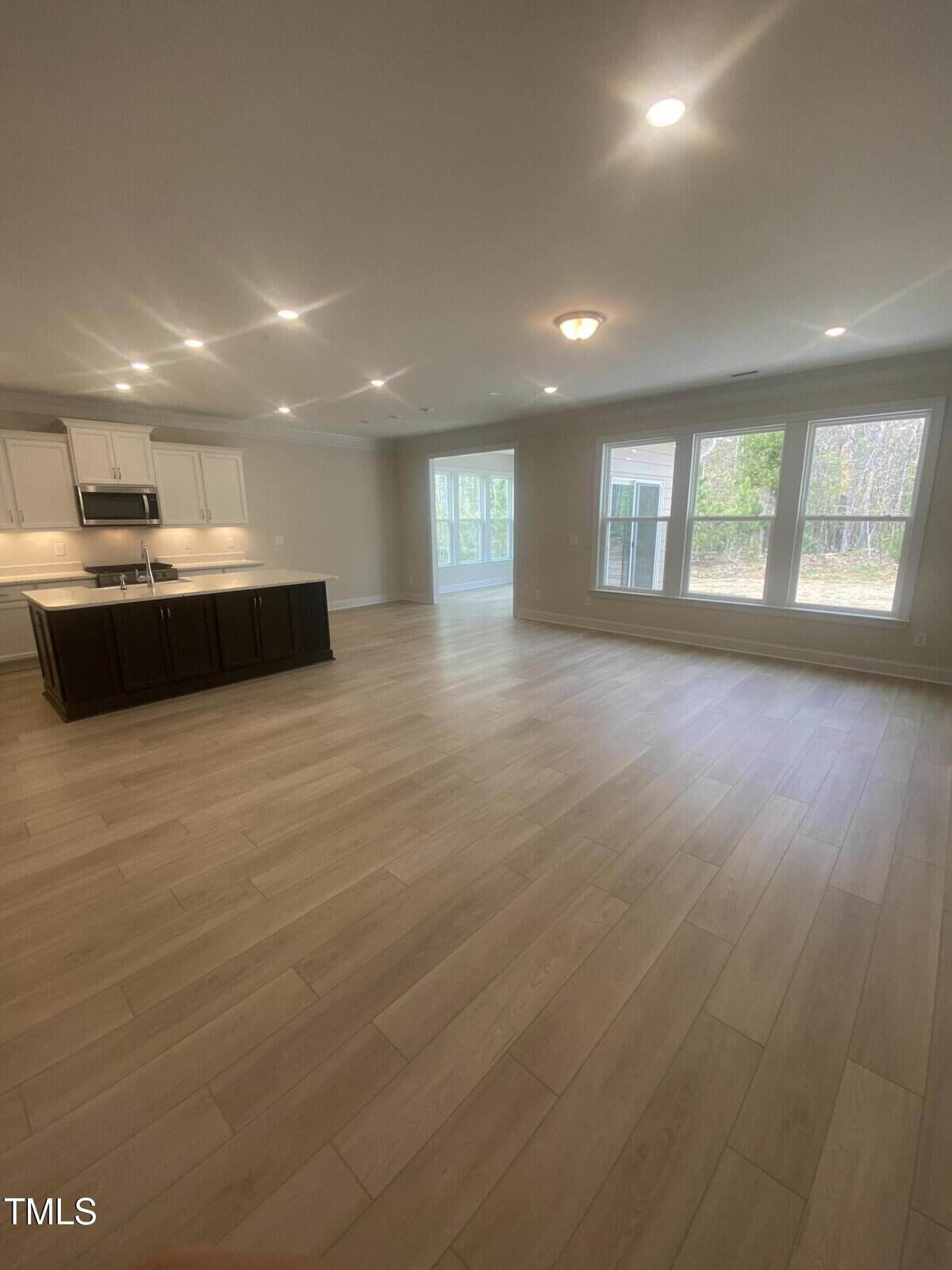 Spacious open-concept living area with light wood flooring, large windows, and recessed lighting. Kitchen features white cabinets, stainless steel appliances, and a dark island. Glass doors lead to an outdoor area.
