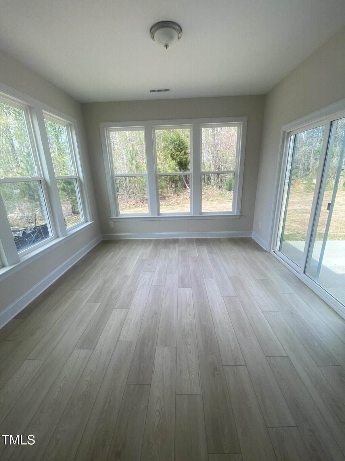 A bright, empty room with light wood flooring, beige walls, and multiple large windows on two sides. A glass sliding door on the right wall leads outside, and trees are visible through the windows.