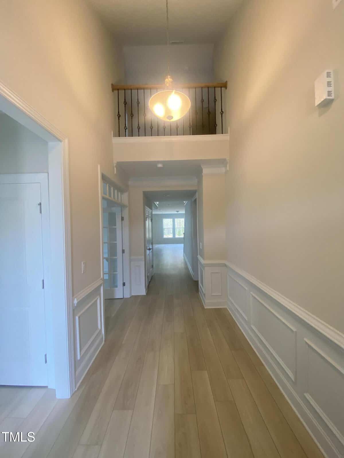 A hallway with light wood flooring, white wainscoting, and beige walls. A light fixture hangs from the ceiling, and there is an open balcony above. Doorways and a bright room are visible at the end of the hallway.