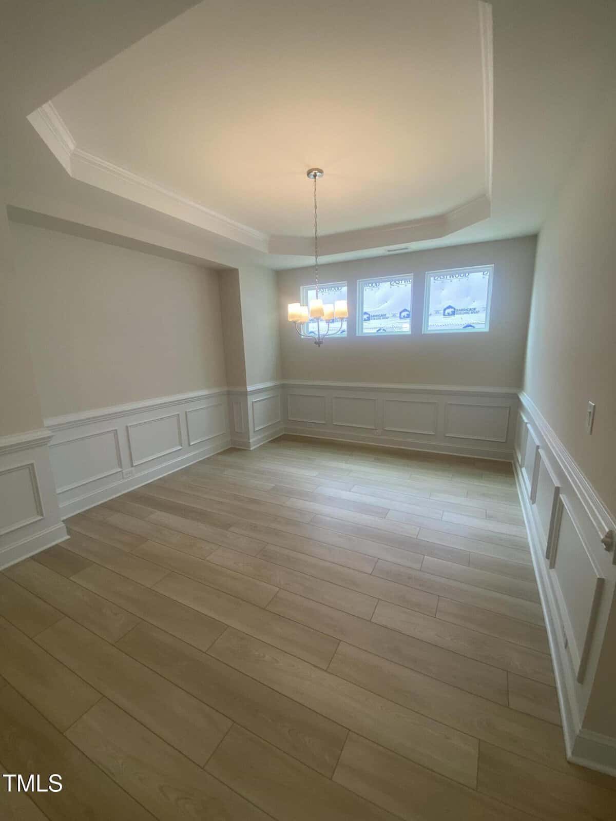 Empty room with light wood flooring, beige walls, white wainscoting, a tray ceiling, a modern hanging light fixture, and three rectangular windows letting in natural light.