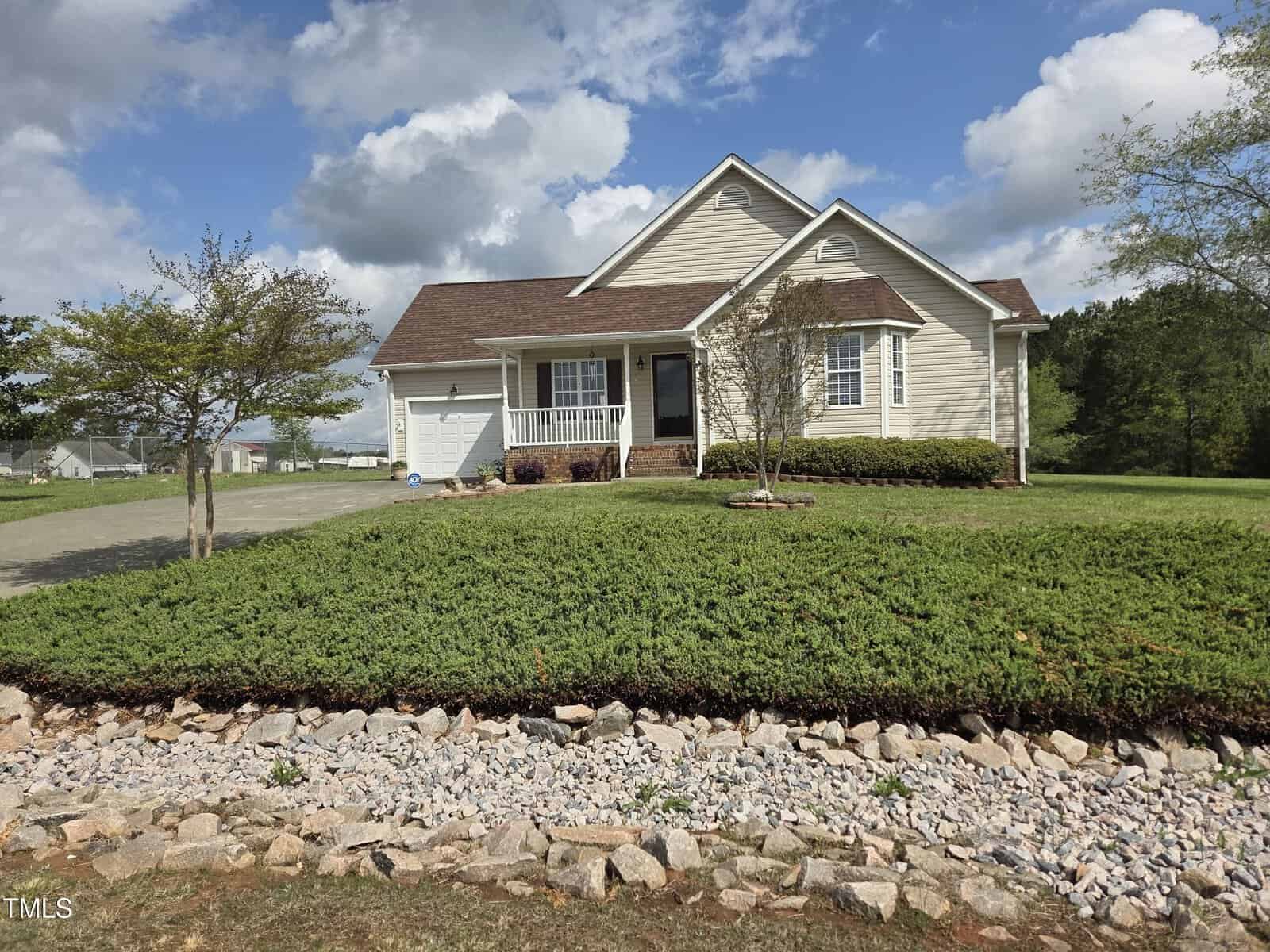 A single-story house at 46 Grain Lane features a front porch and gable roof, all embraced by Henderson's charm. A well-maintained lawn surrounds it, while a stone-bordered garden with greenery graces the foreground. The partly cloudy sky casts gentle shadows across the property.
