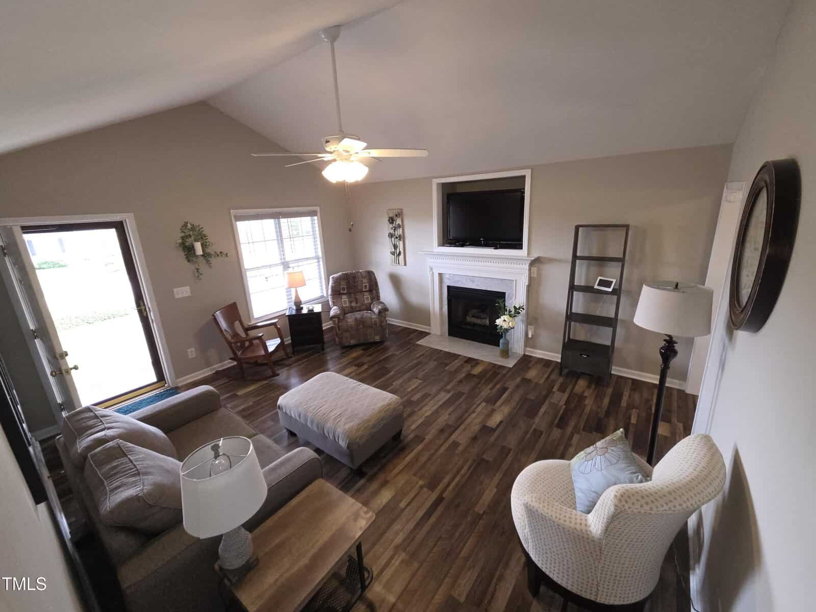 A cozy living room with hardwood floors features a sofa, an armchair, and a recliner chair. A fireplace topped with a TV is centered on the far wall. Near the window are a floor lamp and a small table. A ceiling fan with a light is above.