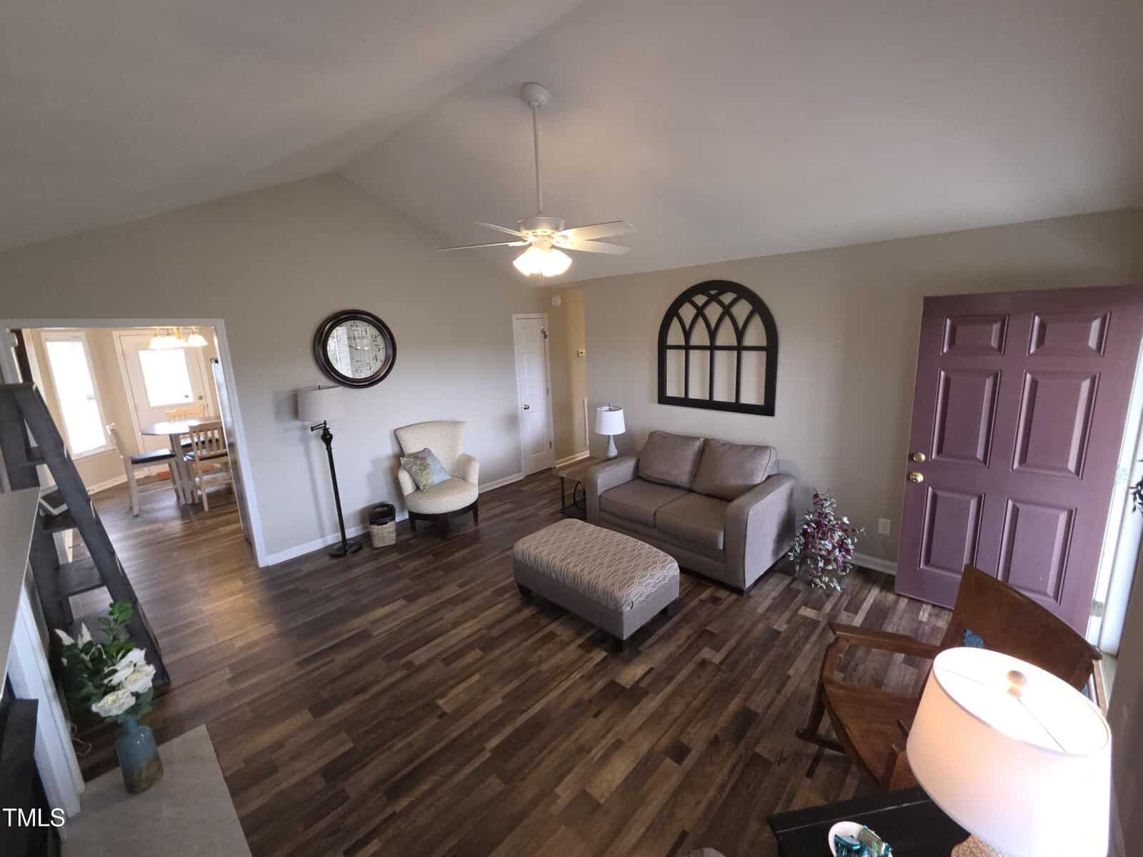 Living room with wood flooring, a sofa, ottoman, and a rocking chair. A ceiling fan hangs from the vaulted ceiling. A round mirror and window-shaped decor adorn the walls. A dining area is visible through an open doorway.