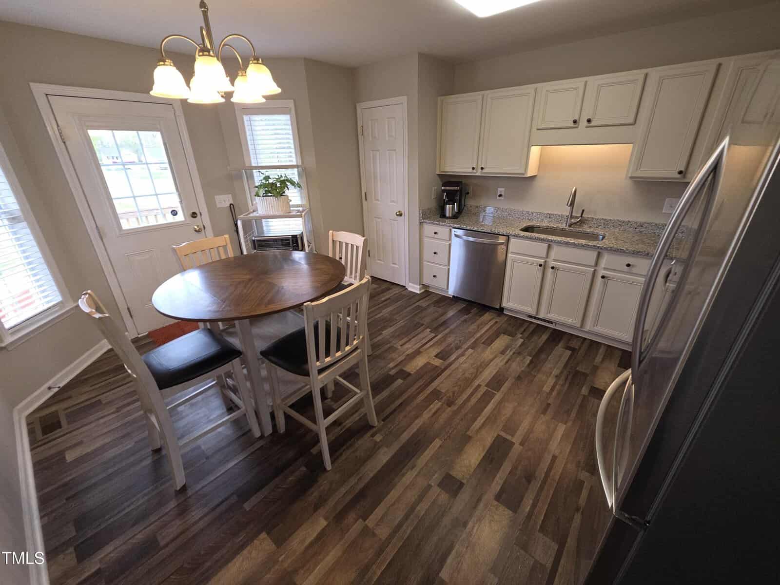 A kitchen with wood-patterned flooring, white cabinets, and stainless steel appliances. A round table with four wooden chairs sits in the center. A door and window are on the left, and a chandelier hangs above the table.