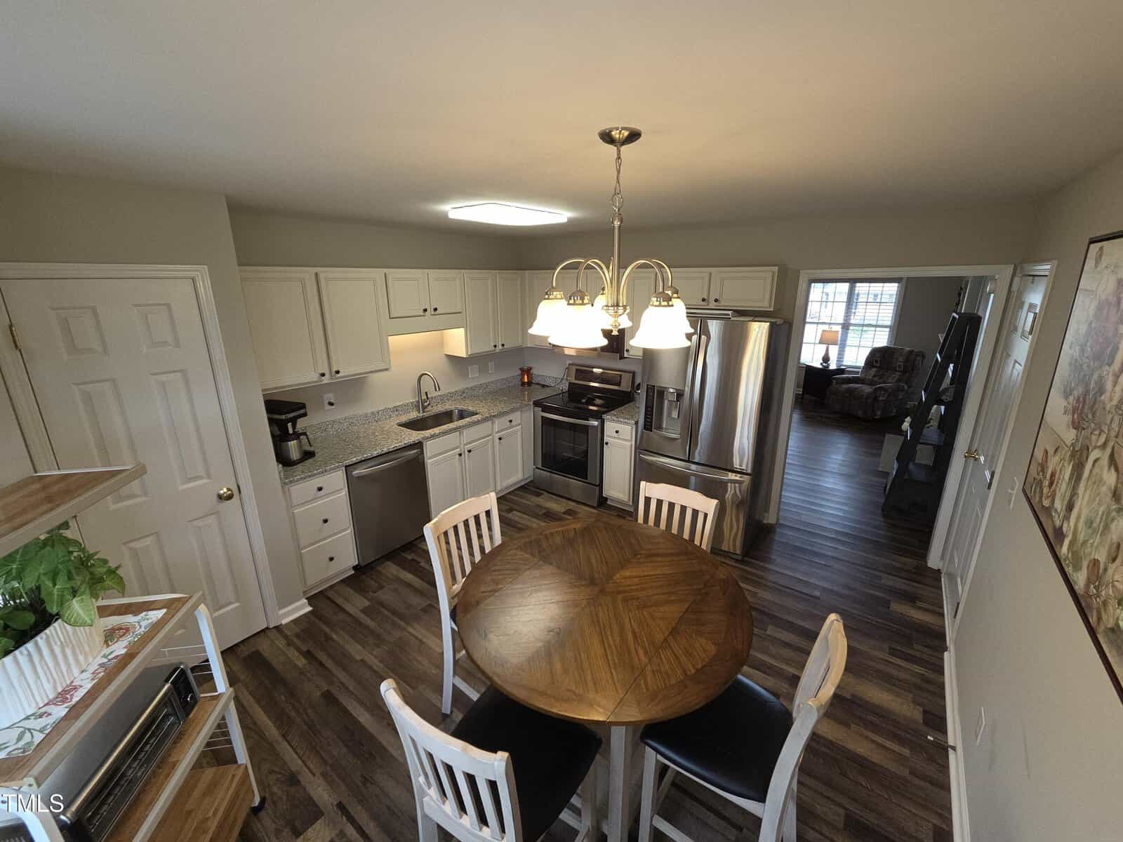 Spacious kitchen with white cabinets, stainless steel appliances, and a wood-topped round table with four chairs. Pendant light hangs above the table. Hardwood flooring and adjacent living area with a couch are visible. Large windows provide natural light.