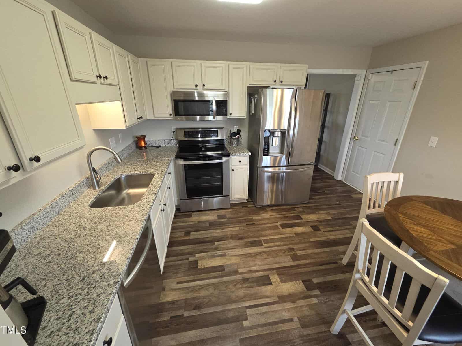 A kitchen with white cabinets, granite countertops, and stainless steel appliances including a microwave, stove, and refrigerator. There is a round wooden table with two chairs on a hardwood floor.