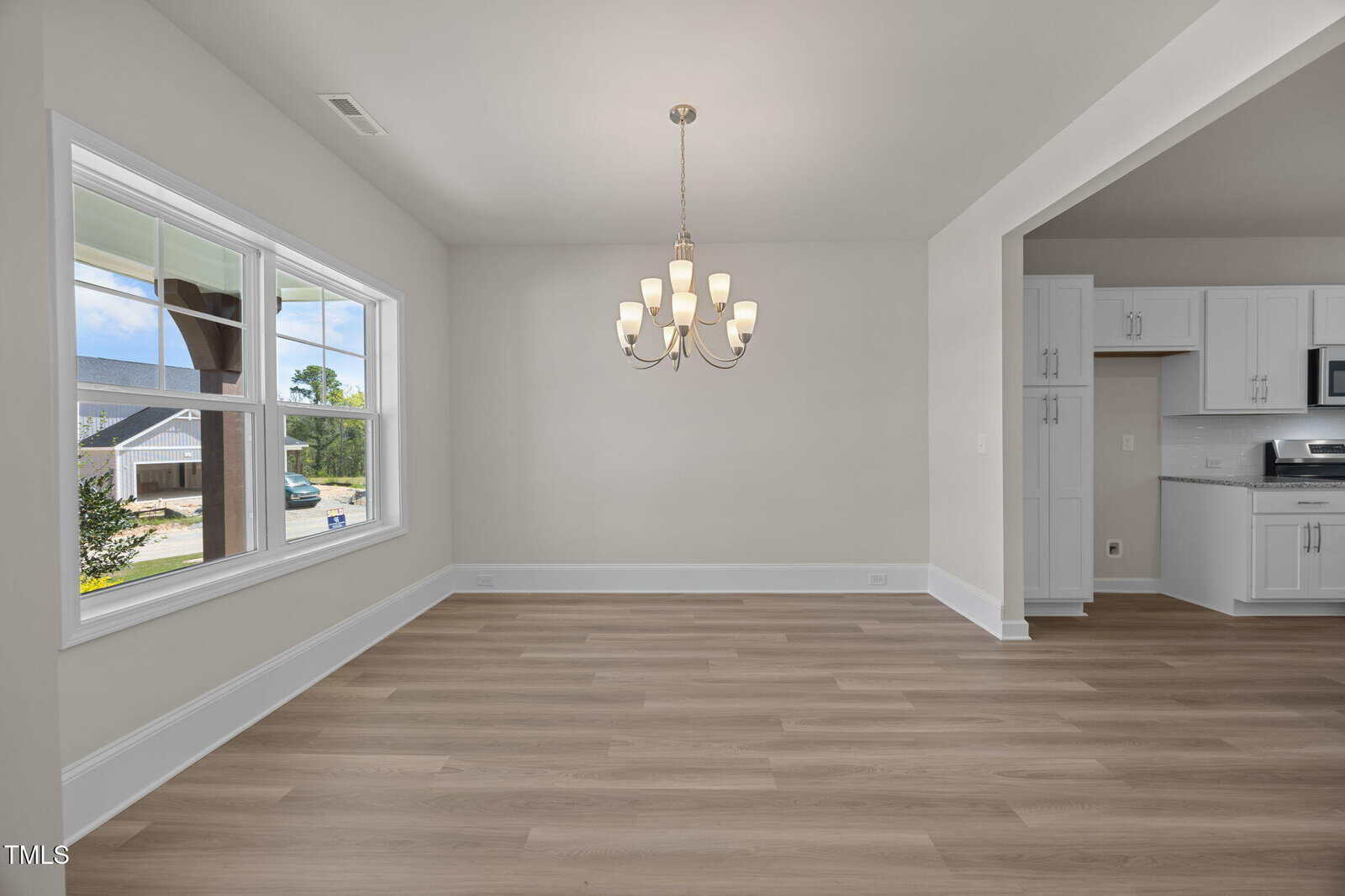 A bright, empty dining area with light wood floors, white walls, a large window, a modern chandelier, and an open view into a white kitchen with stainless steel appliances.