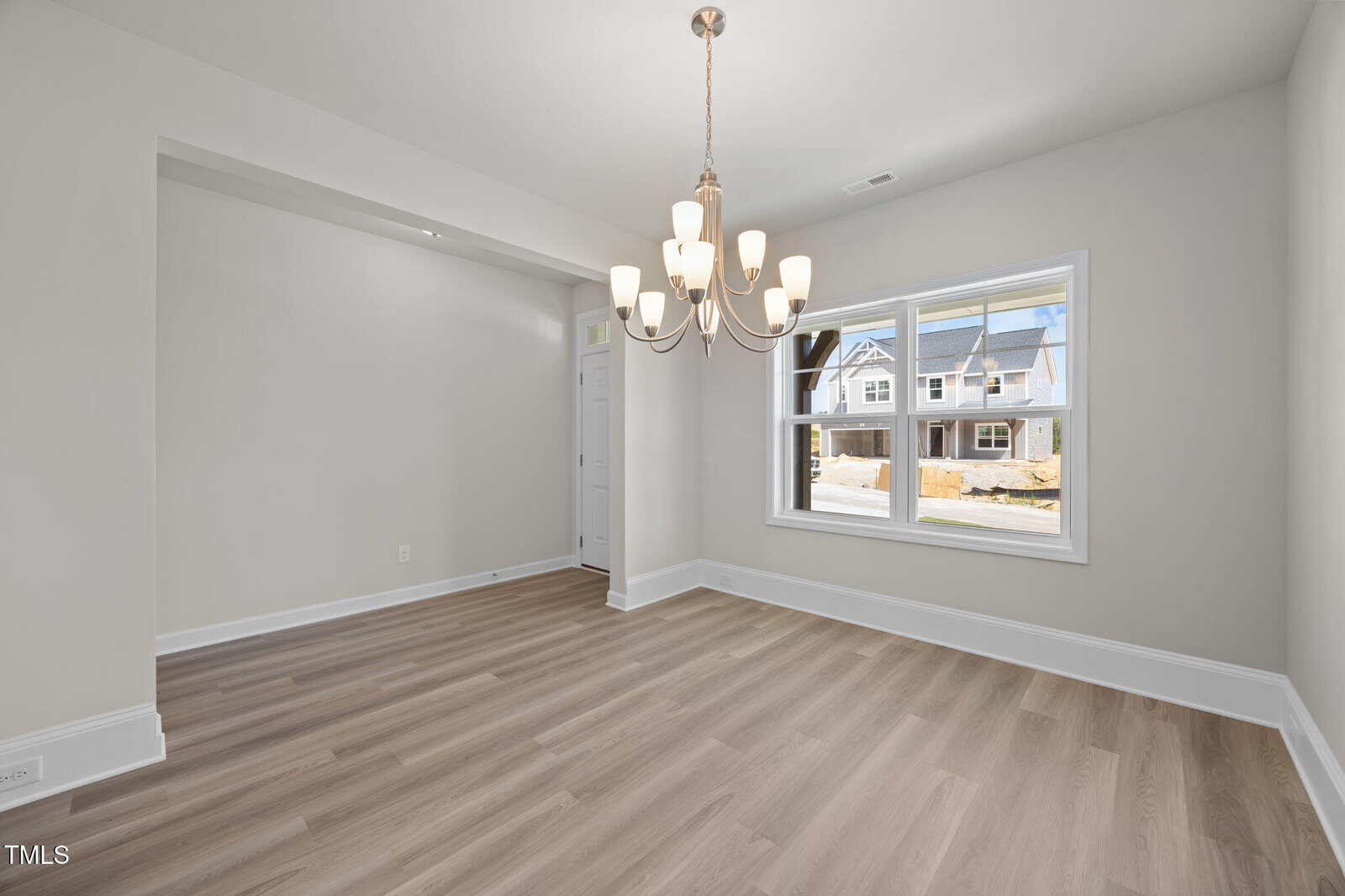 A bright, empty room with light wood flooring, neutral-colored walls, a window showing houses outside, and a modern chandelier hanging from the ceiling.