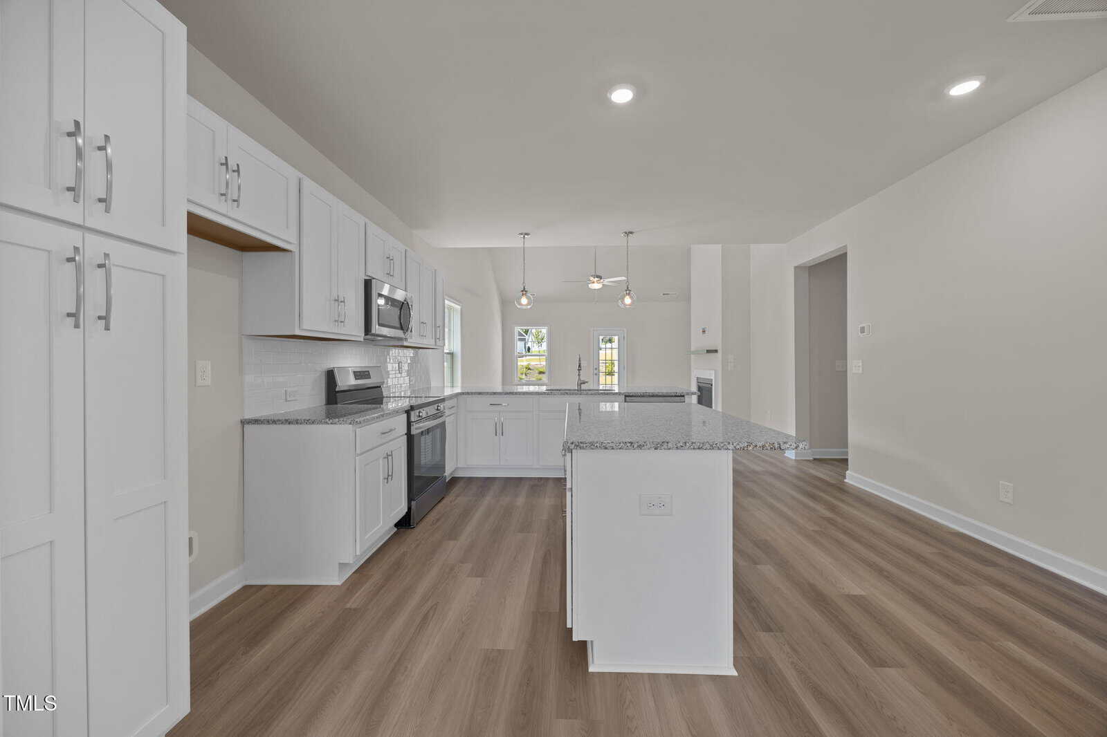 A modern kitchen with white cabinets, stainless steel appliances, granite countertops, and light wood flooring. Pendant lights hang above a central island, and large windows provide natural light.