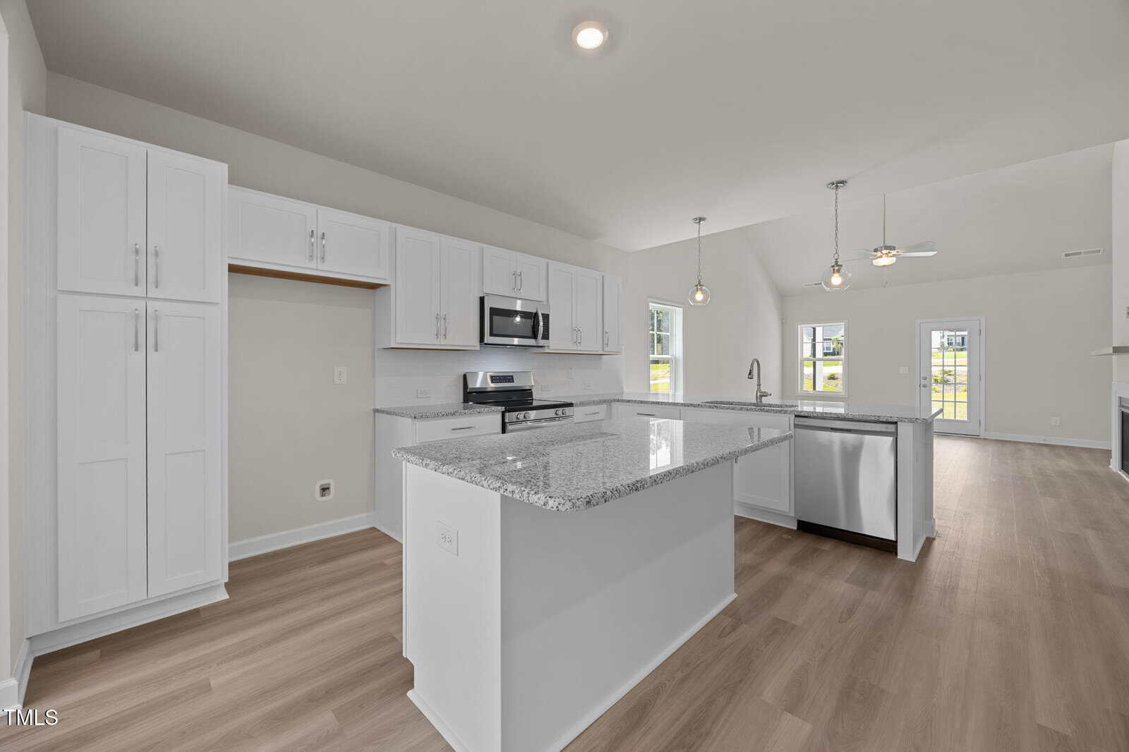 Modern kitchen with white cabinets, stainless steel appliances, granite countertops, a large island with a sink and dishwasher, light wood flooring, and an open layout leading to a bright living area with ceiling fan.