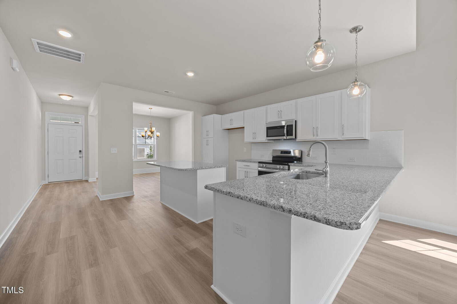 Bright, modern kitchen with white cabinets, stainless steel appliances, two granite islands, pendant lights, and light wood flooring; open layout leads to dining area and entryway in the background.