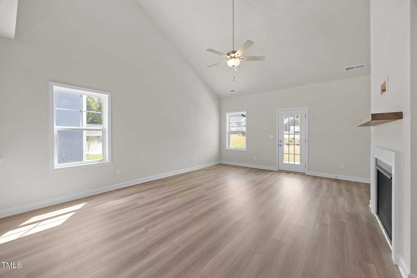 A bright, empty living room with vaulted ceilings, light wood flooring, two windows, a ceiling fan with light, a door to the backyard, and a white fireplace on the right wall. Sunlight streams through the windows.