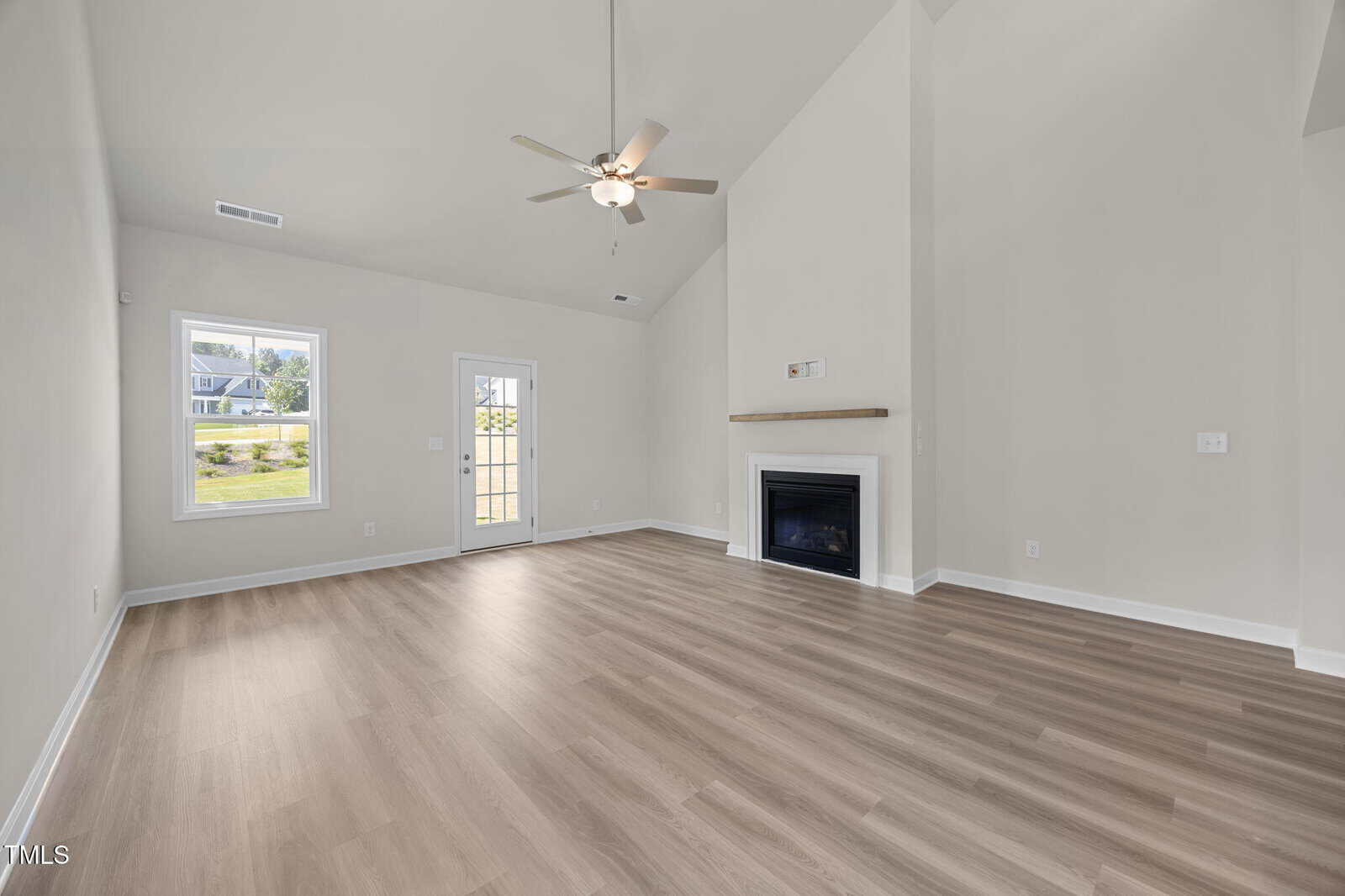 Spacious, empty living room with light wood flooring, high vaulted ceiling, ceiling fan, white walls, a fireplace with a small mantle, and large windows letting in natural light. A glass door leads outside.