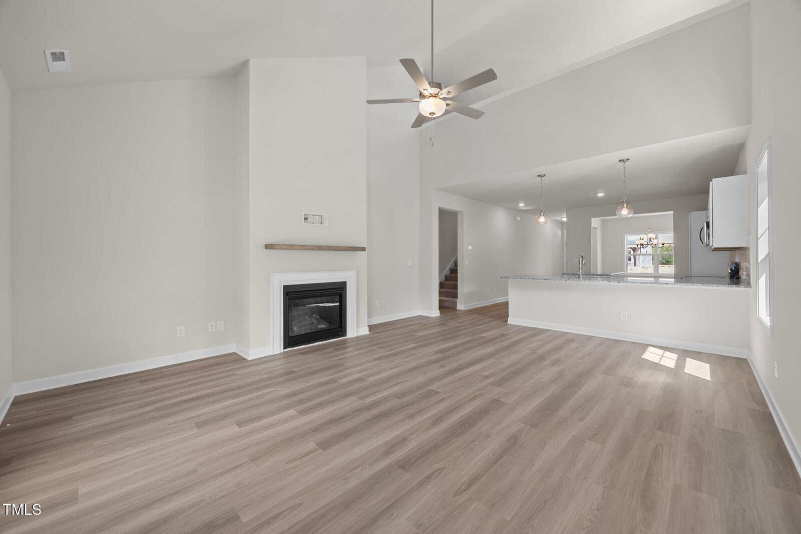 Spacious modern living area with light wood floors, a ceiling fan, a white fireplace, and an open kitchen with pendant lights and a large breakfast bar.