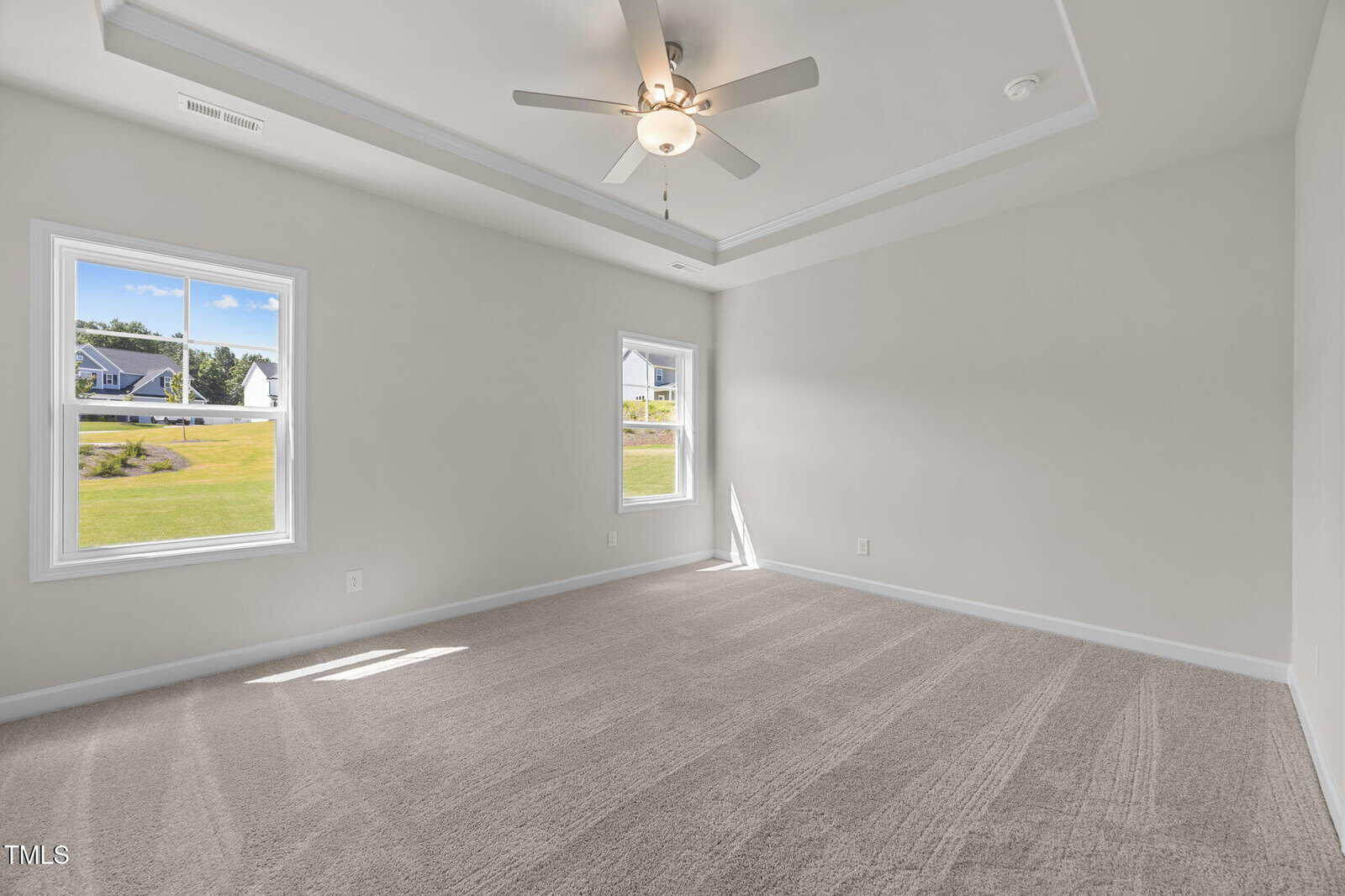 Bright, empty bedroom with light gray walls, beige carpet, tray ceiling, ceiling fan with lights, and two windows letting in natural daylight, offering a view of a green lawn and houses outside.