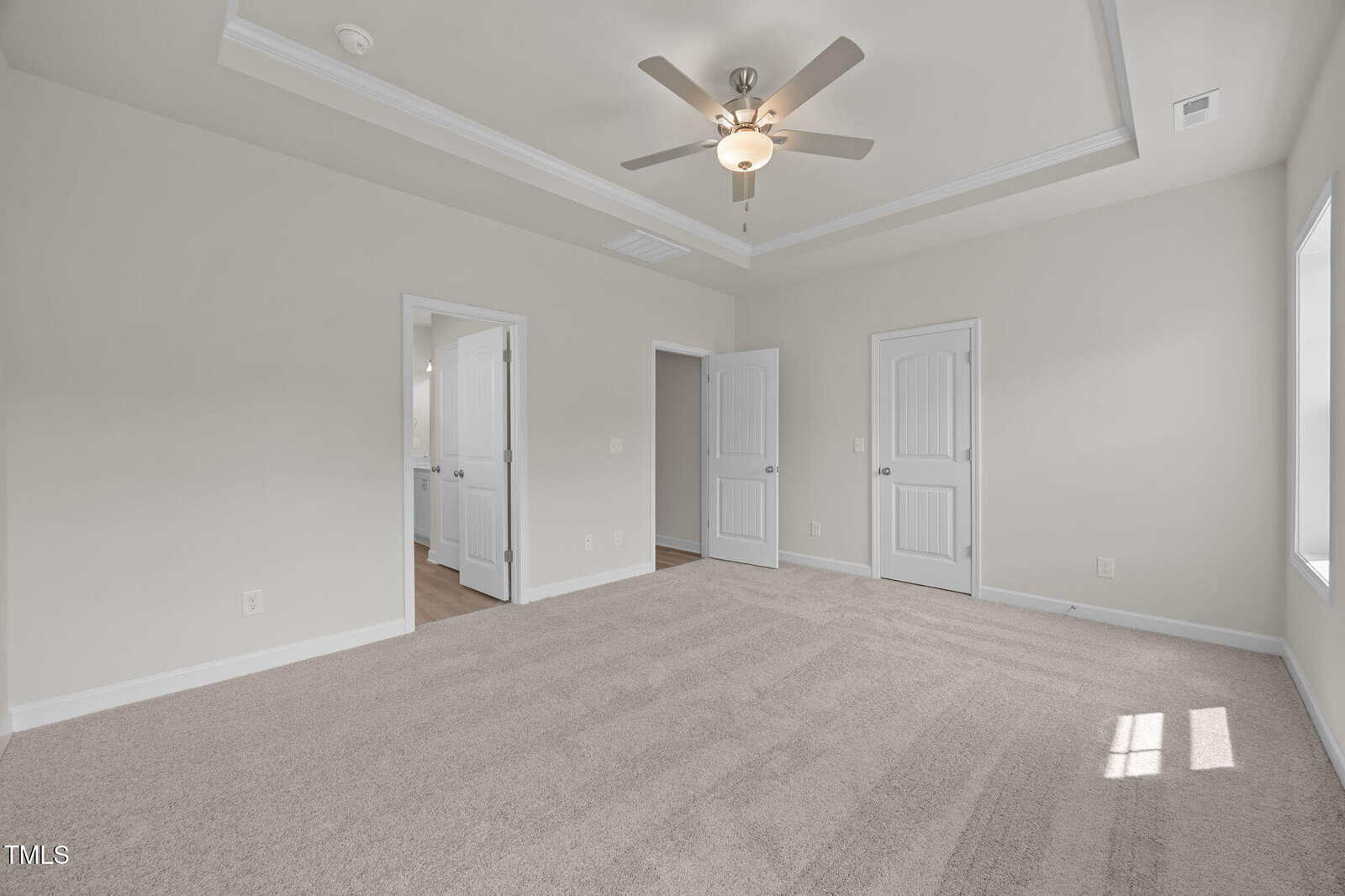 Bright, empty carpeted bedroom with light gray walls, ceiling fan, tray ceiling, two white doors, one open to a bathroom, and sunlight streaming through a window on the right.
