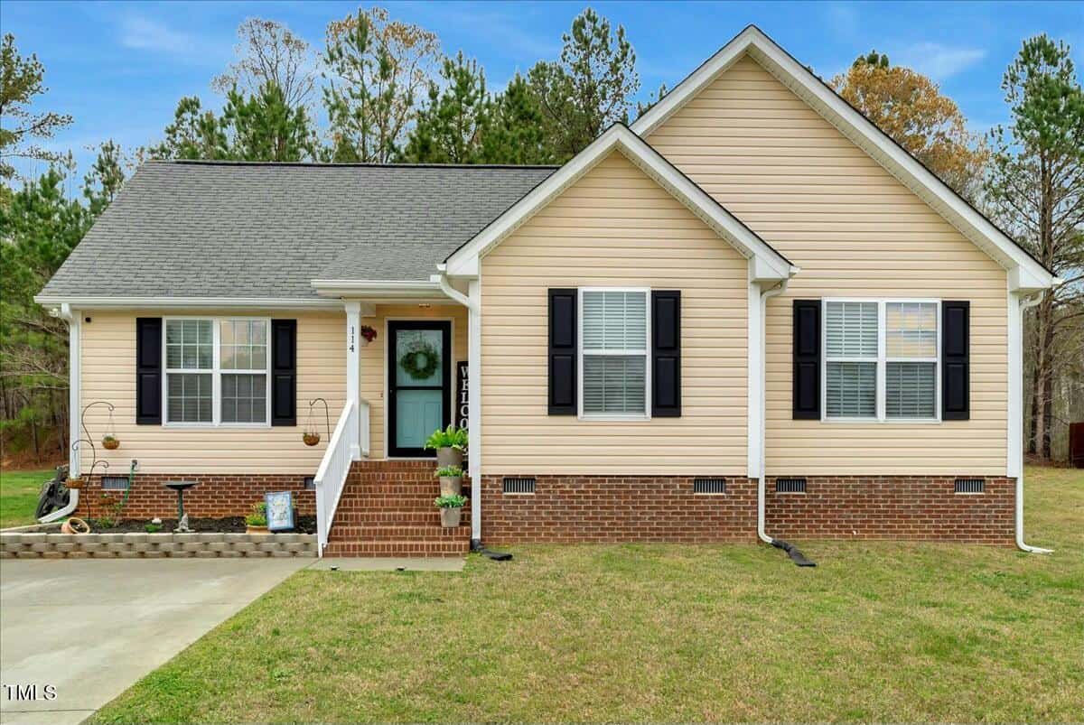 Located at 114 Carriage Hill Drive, this charming single-story beige house features black shutters and a gray roof. A white-railed front porch with potted plants welcomes you, while a concrete driveway stems gracefully from the steps, all surrounded by a lush grassy lawn and idyllic trees.