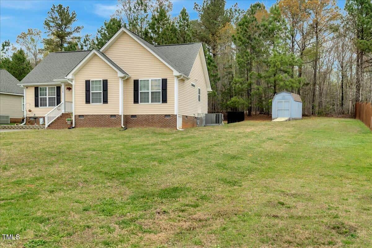 A suburban house with beige siding and a dark roof is located on a large grass lawn. There are trees in the background and a white shed on the right side. The house has small steps leading to the front door.