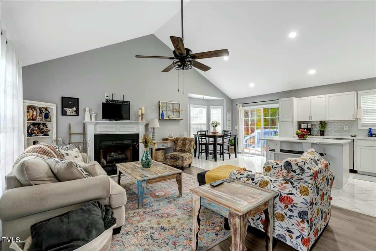 Spacious, well-lit living room with high ceiling and ceiling fan. Features a fireplace, patterned sofa, armchairs, coffee tables, and a colorful rug. Open kitchen and dining area with white cabinets and large windows in the background.