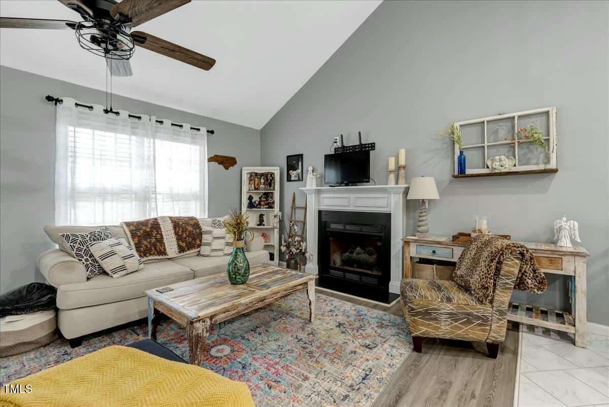 A living room with a beige sofa, patterned throw pillows, and a wooden coffee table. There's a fireplace with decor above, and various art pieces and shelves around. A colorful rug covers the floor, and a ceiling fan is above. The walls are gray.