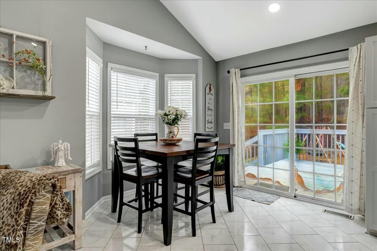 A bright dining area features a dark wooden table with four matching chairs. Large windows and sliding glass doors offer views of an outdoor deck and pool. Neutral-toned walls, decorative items, and a leopard-print throw complete the decor.