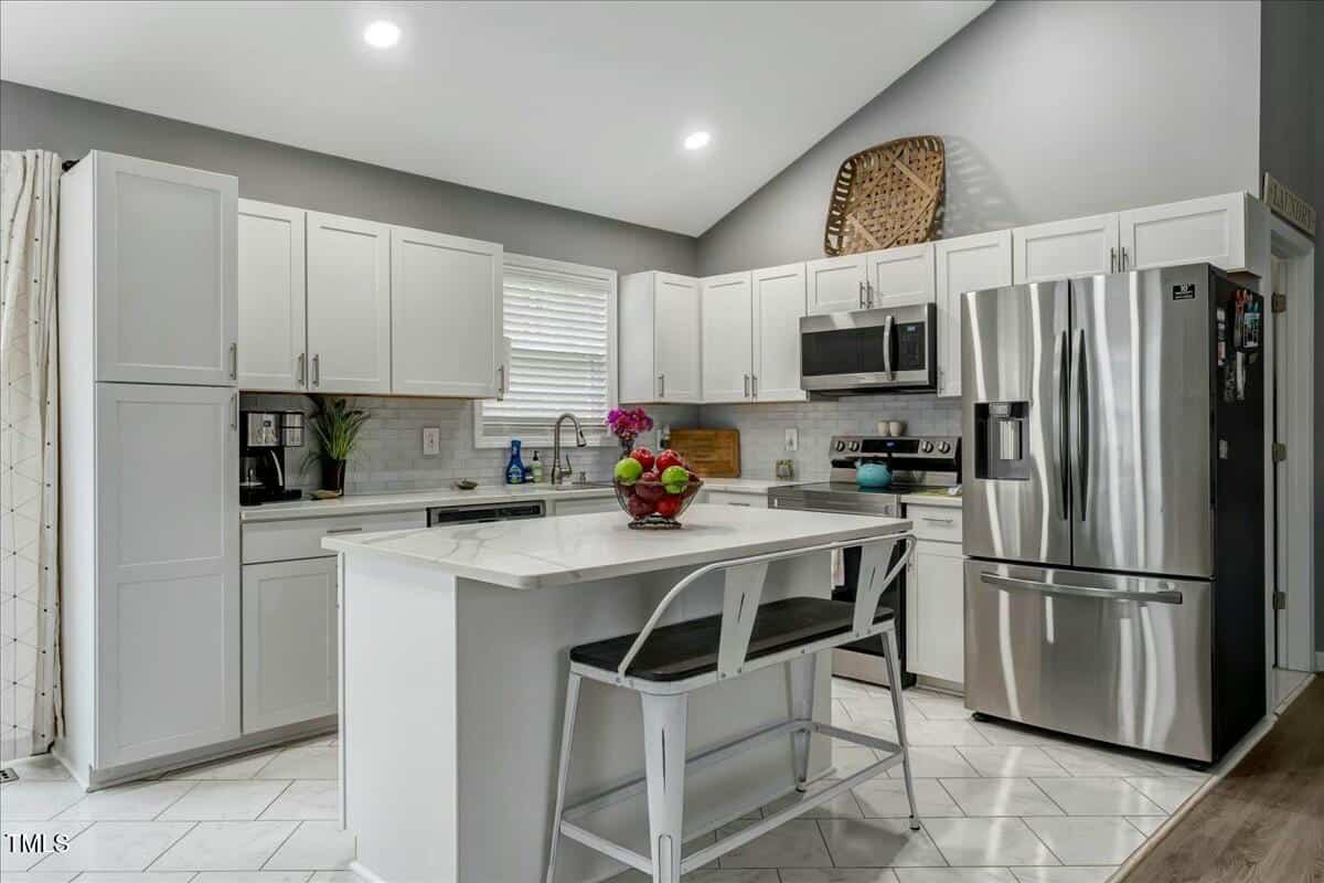 Modern kitchen with white cabinets, stainless steel appliances, and a marble-topped island. Two high-backed metal chairs are at the island, with a fruit bowl on top. Gray walls and a woven basket above the cabinets add decor.