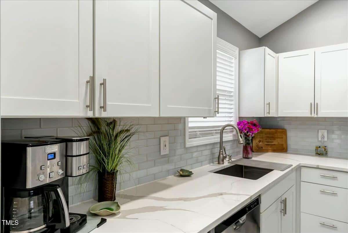 Modern kitchen with white cabinets and marble countertop. Features a stainless steel sink, a coffee maker, cutting board, and a potted plant. Natural light enters through a window with blinds. Gray subway tiles line the backsplash.