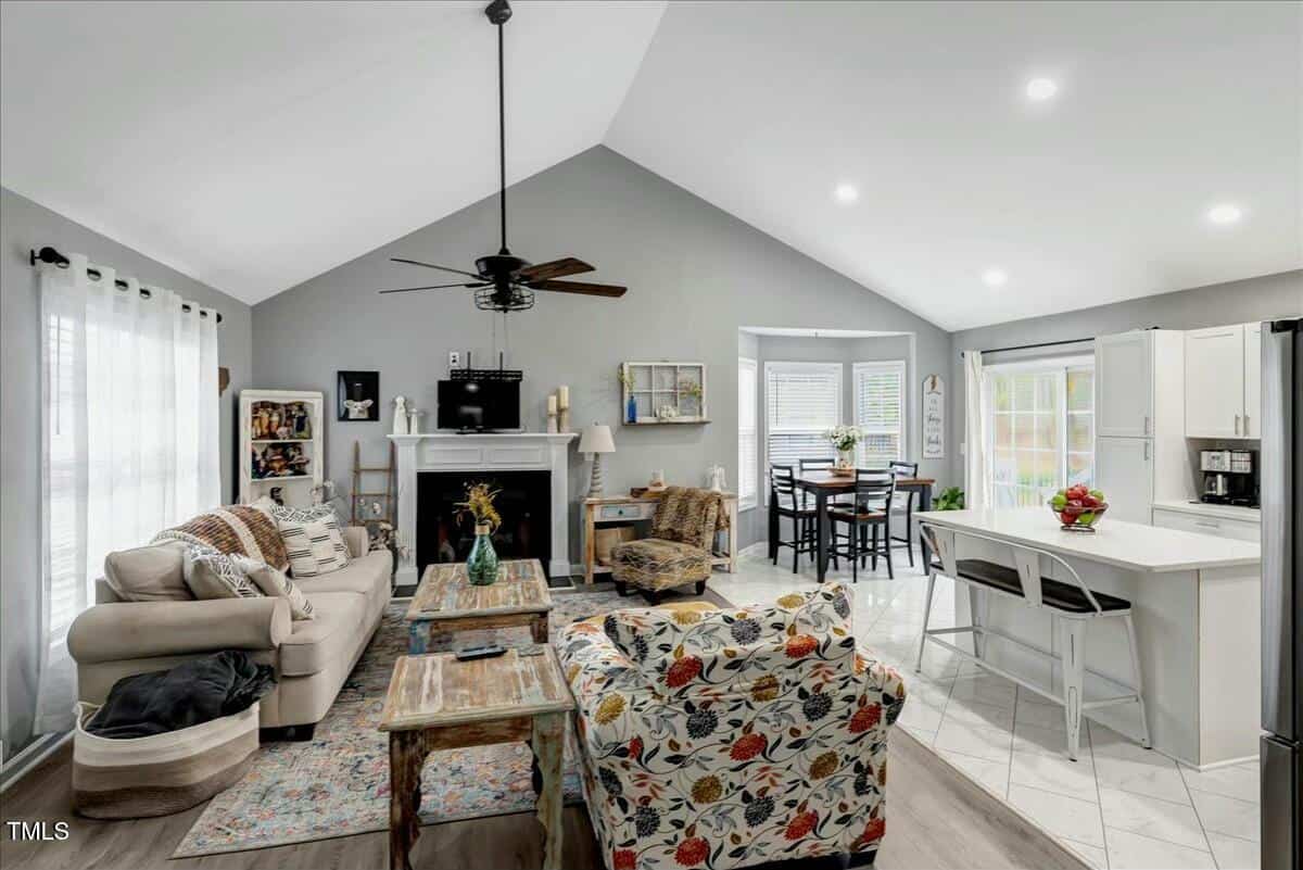 Open-concept living room and kitchen with vaulted ceiling. Features include a beige sofa, floral-patterned chair, fireplace with TV, dining table, and kitchen island with bar stools. Light gray walls and large windows provide ample natural light.