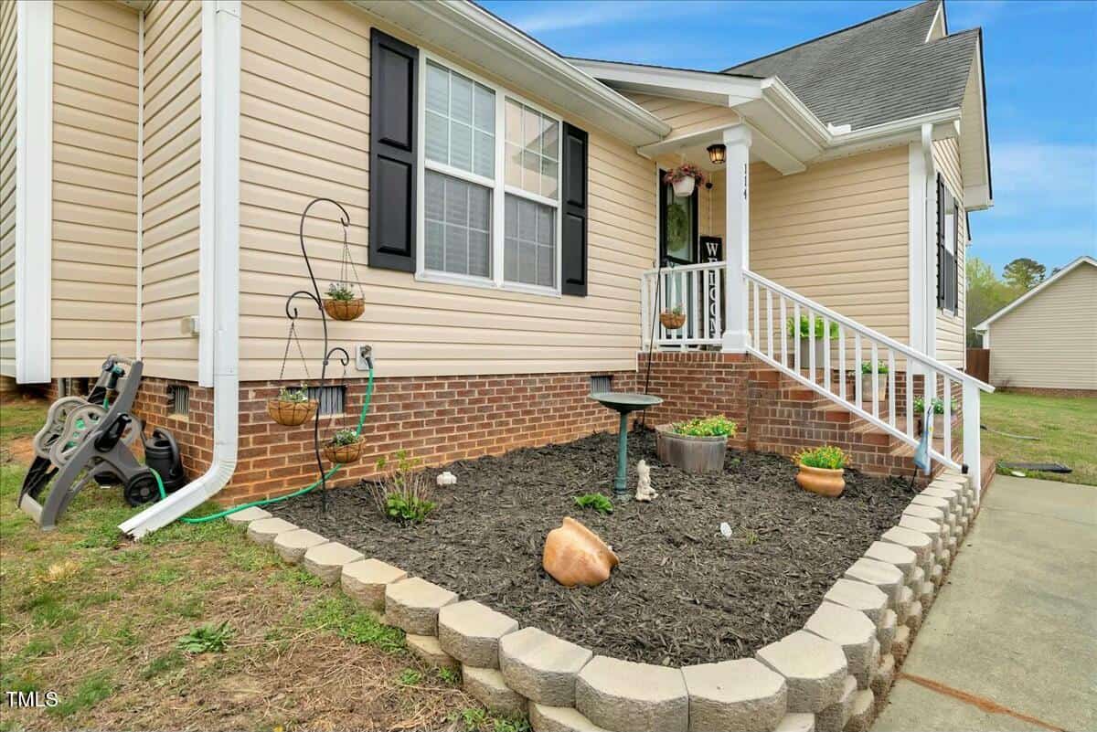The image shows the corner of a beige house with white trim and brick foundation. There is a small garden bed bordered by stones, containing a birdbath, potted plants, and garden decor. A hose is coiled nearby, and a white railing leads to the entrance.