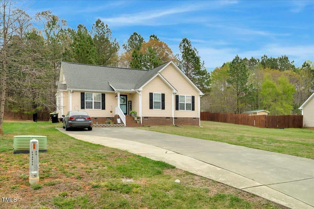 A single-story beige house with a dark gray roof and front porch is visible. A black car is parked in the driveway. The house is surrounded by a grassy yard and trees are in the background. A mailbox and utility box are near the driveway.