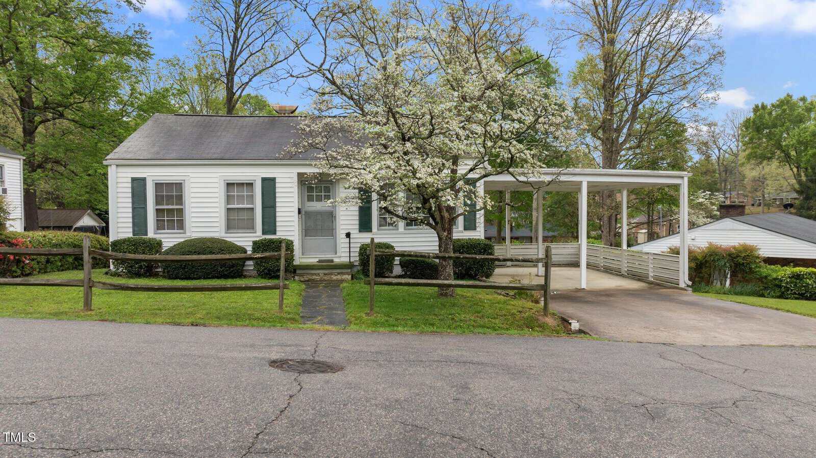 A small white single-story house at 1631 Sunset Avenue features a covered carport on the right. With green shutters, a front path, and a blossoming tree in the yard, this charming home is nestled on a slightly elevated grassy area in Henderson beside a paved street.