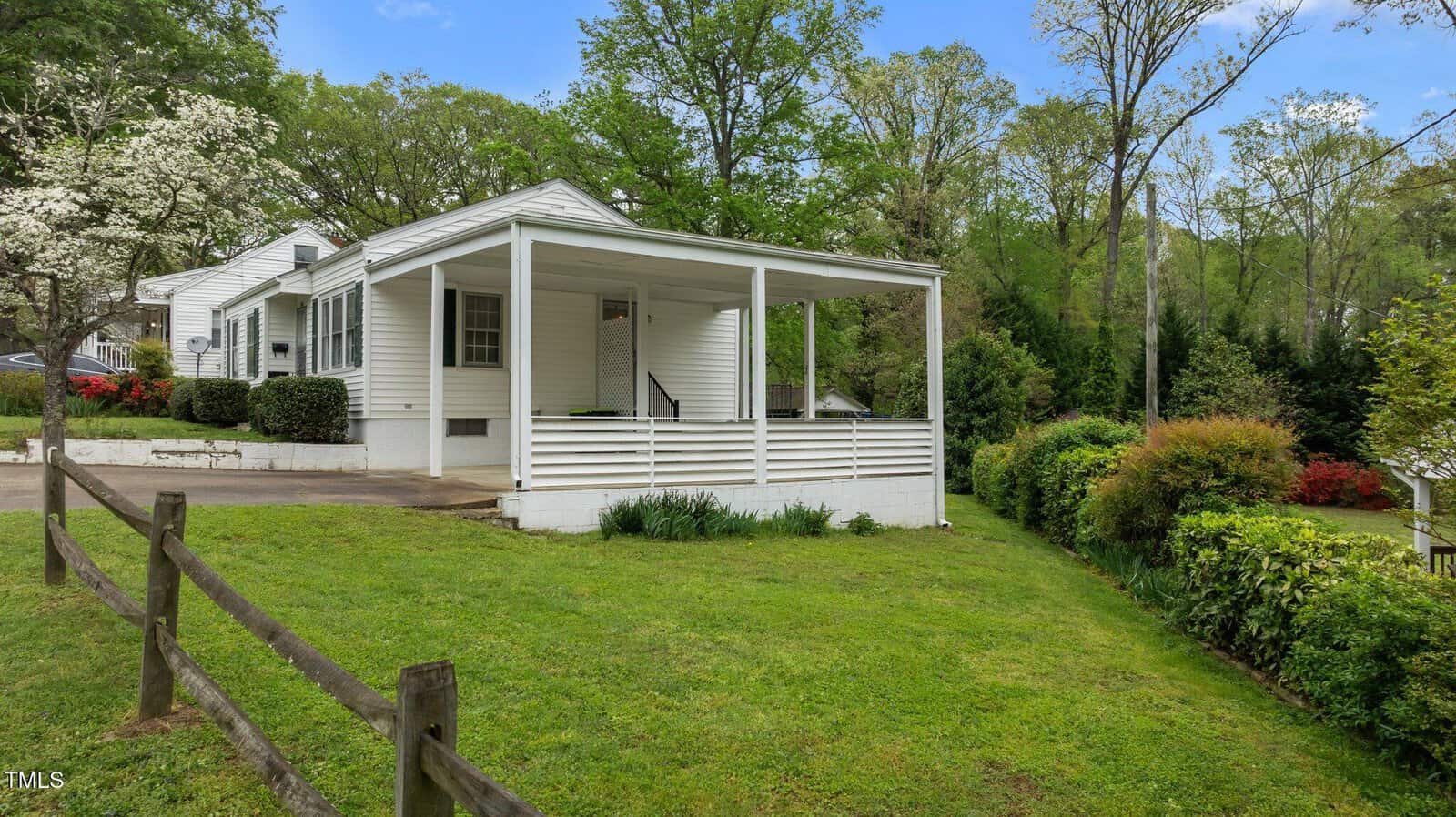 A white, single-story house with a covered porch is surrounded by greenery and a neatly trimmed lawn. A wooden fence borders the property, and trees and shrubs provide a natural backdrop. The sky is clear with scattered clouds.