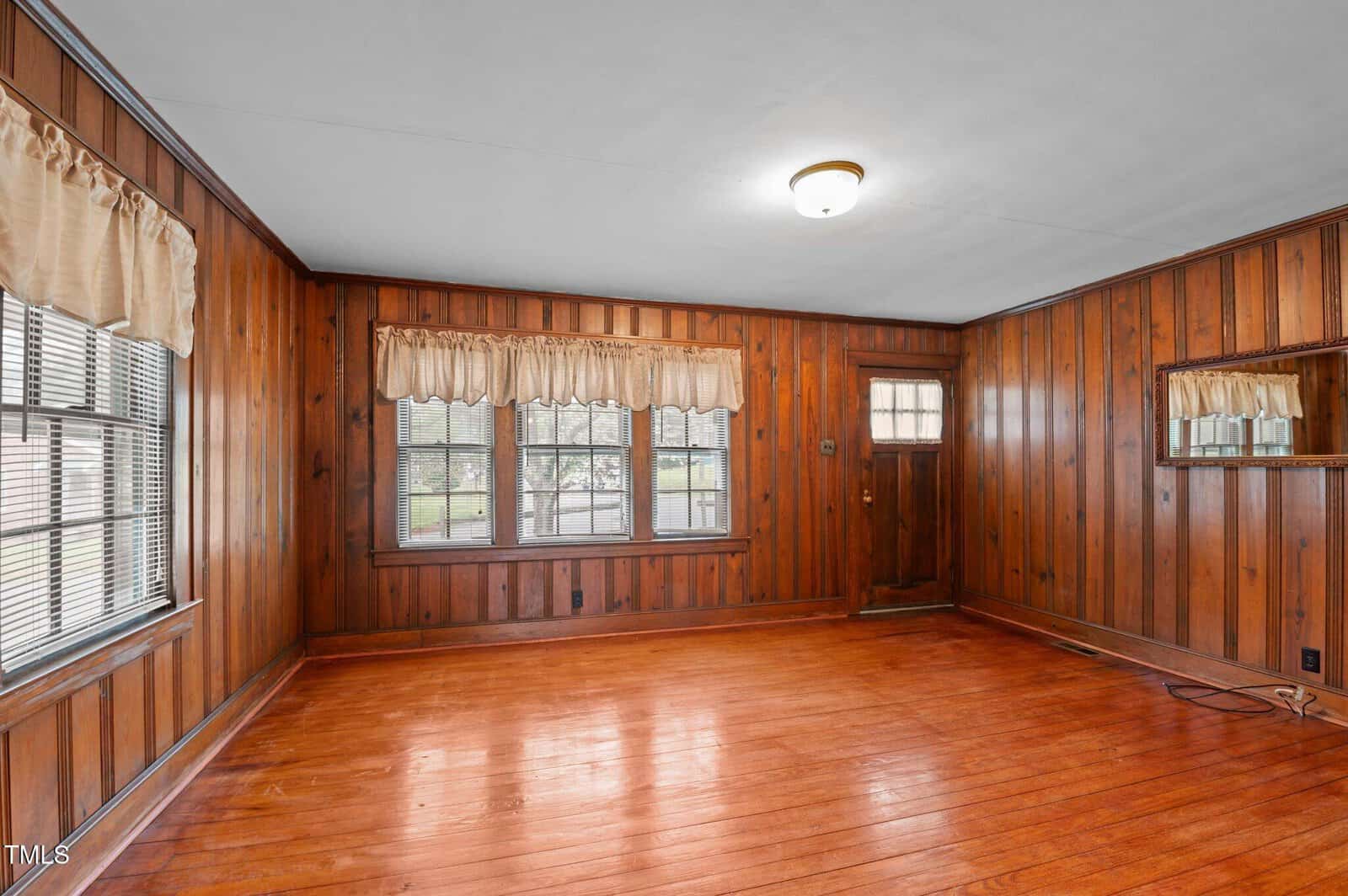 A room with wooden walls and flooring features three large windows with beige curtains. There is a wooden door with a window and a small wall-mounted mirror. A ceiling light illuminates the space.