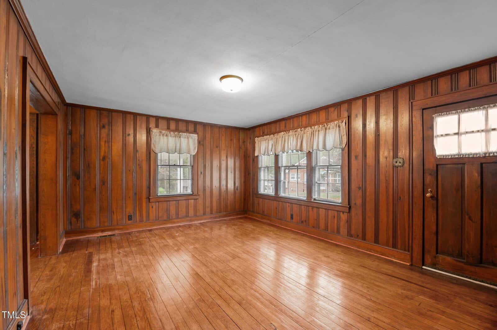 A wood-paneled room with a wooden floor features two windows with light-colored curtains and a wooden door with a frosted glass panel. A ceiling light fixture is centered on the white ceiling.