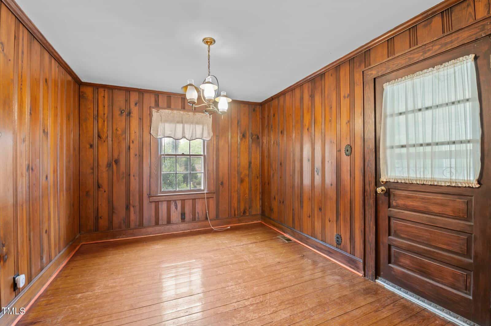 A room with wooden panel walls and a wooden floor. It has a window with a curtain and a door with a curtain above the knob. The ceiling features a hanging light fixture with three bulbs. The room is empty and well-lit.