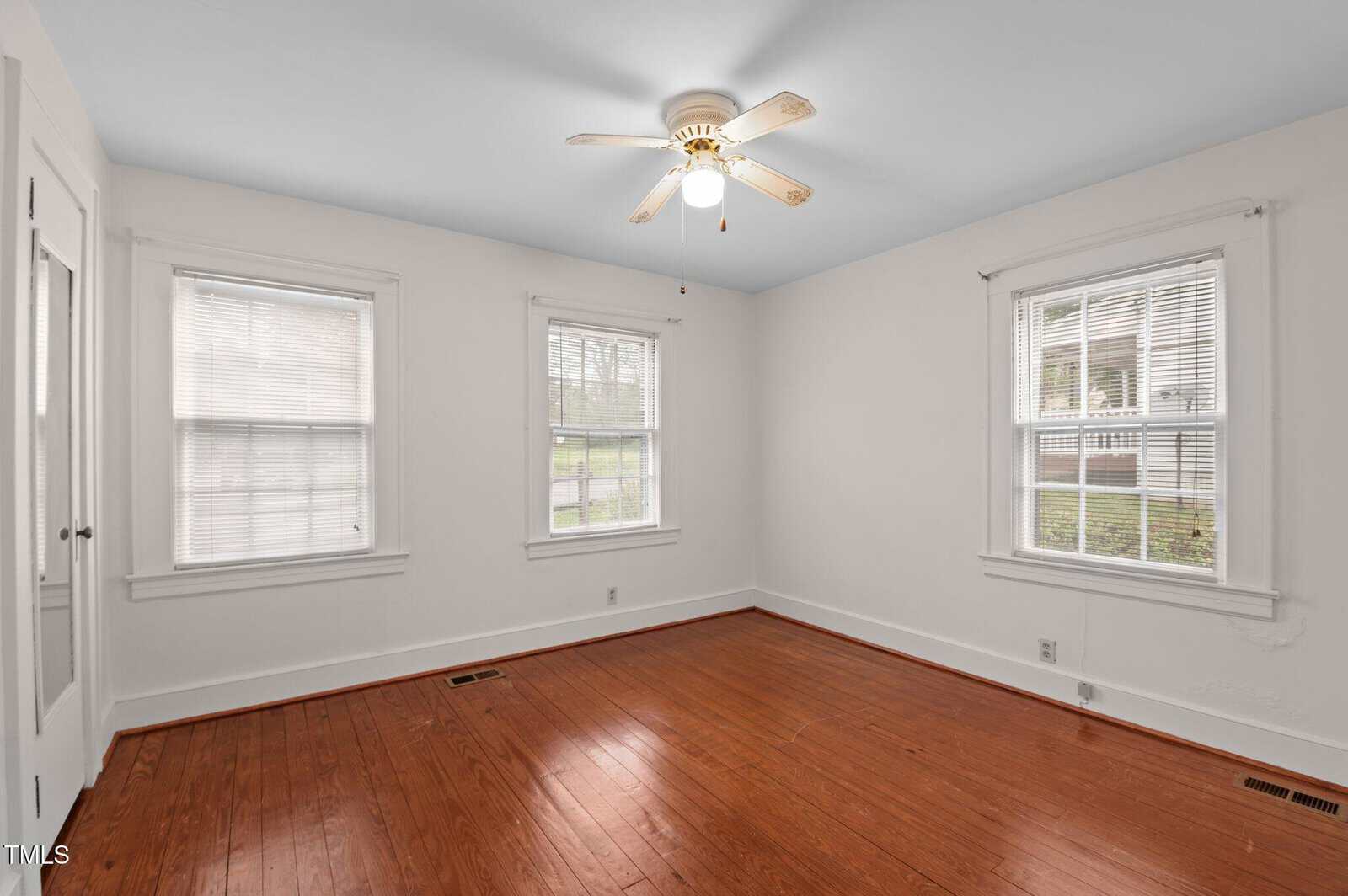 An empty room with white walls and a ceiling fan. The floor is hardwood, and there are two windows with blinds on the right wall. Another window is on the left wall. A vent is visible on the floor, and a closed door is on the far left.