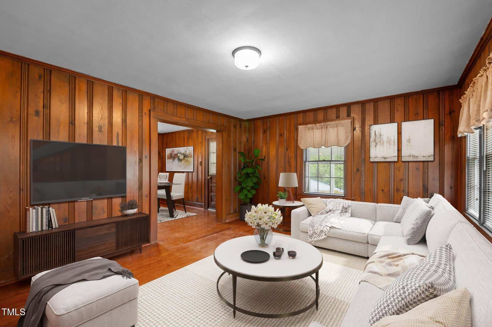 A cozy living room with wooden paneled walls, a ceiling light, and a large window. It features a beige sectional sofa, a round coffee table with flowers, a TV on a stand, and an open doorway leading to a dining room with artwork on the wall.