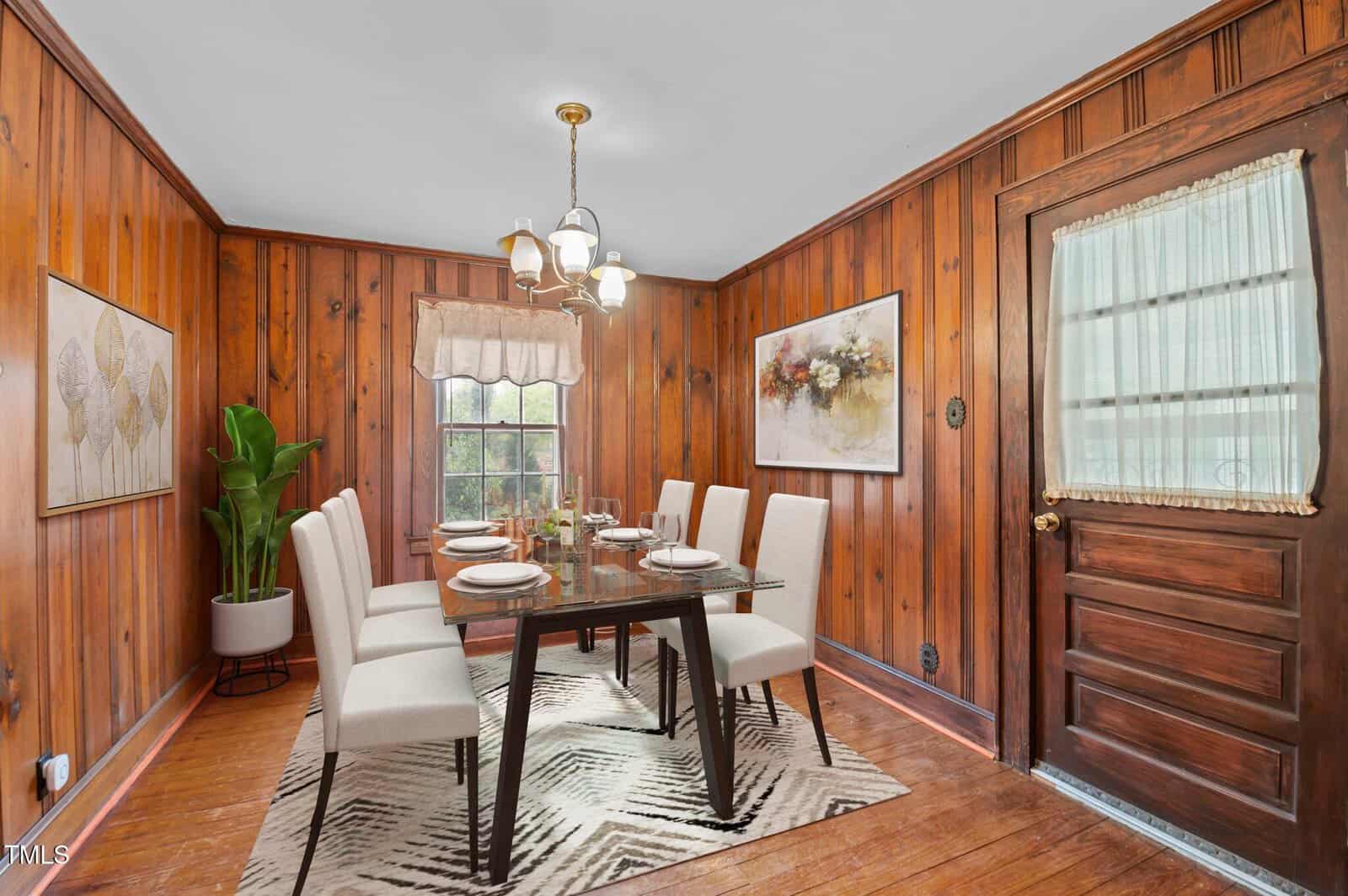 A dining room with wood-paneled walls featuring a rectangular table set for eight. Cream-colored chairs surround the table. A patterned rug is underneath, with wall art, a potted plant, and a wooden door visible. A window has sheer curtains.